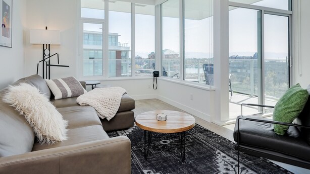 Ein modernes Wohnzimmer mit einem grauen Sofa, einem runden Couchtisch und einem schwarzen Ledersessel. Große Fenster bieten Blick auf die Stadtlandschaft.