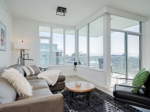 Uma sala de estar moderna com um sofá cinzento, uma mesa de centro redonda e uma poltrona de couro preto. Janelas amplas oferecem vista para a paisagem urbana.