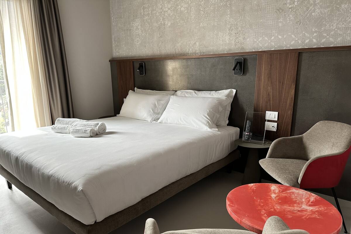 Ein modernes Hotelzimmer mit einem großen Bett, einem Nachttisch mit Fernbedienung, einem rot-grauen Sessel und einem kleinen roten Tisch.