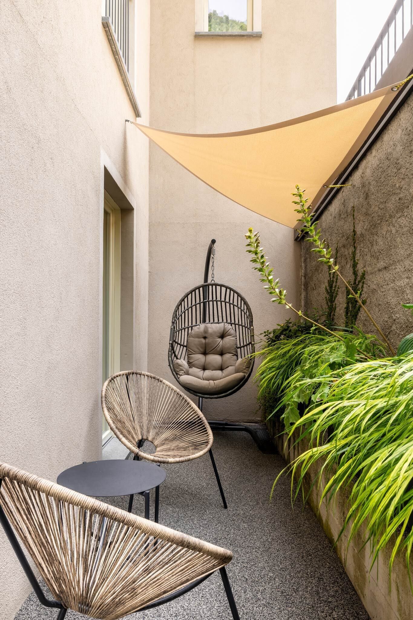 A small outdoor seating area with wicker chairs and a cushioned swing chair, surrounded by plants and a beige shade sail.