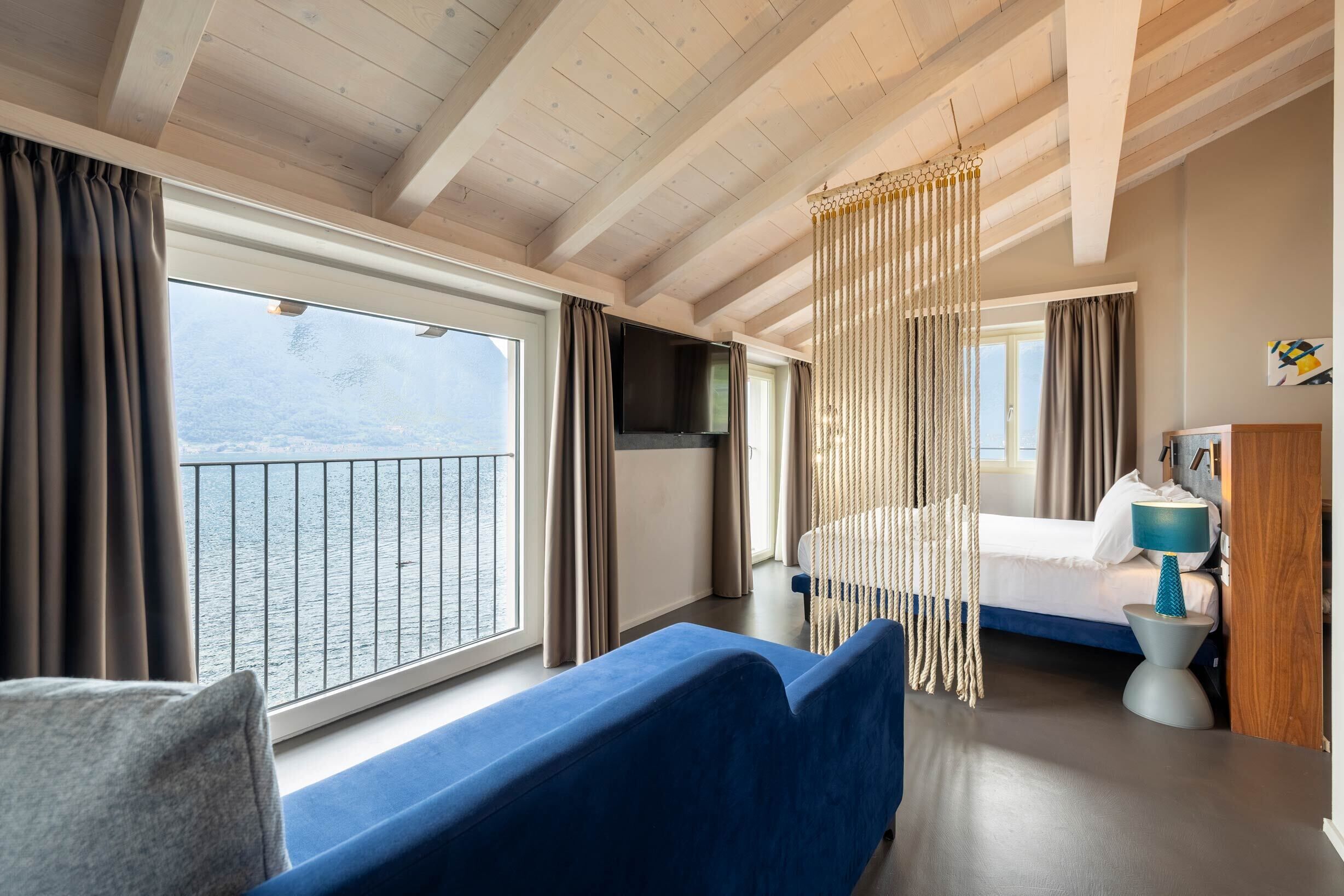 Ein Zimmer mit großem Fenster mit Blick auf ein Gewässer, ein Bett mit blauer Kopfteil, ein blaues Sofa und ein kleiner Nachttisch mit Lampe.