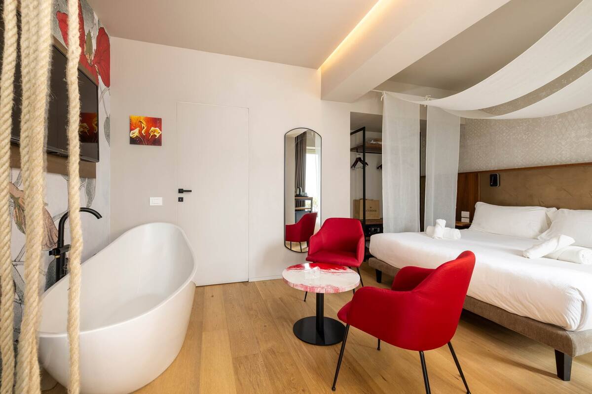 Una habitación de hotel moderna con una bañera grande, una cama con ropa blanca, una silla roja y una pequeña mesa redonda.
