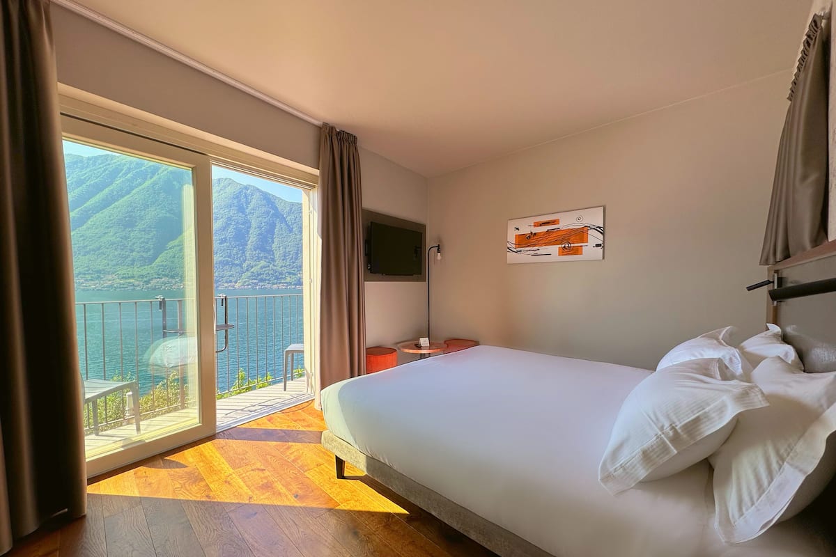 Ein Hotelzimmer mit Bett, Fernseher, einem Gemälde und einem Balkon mit Blick auf Berge und Wasser.