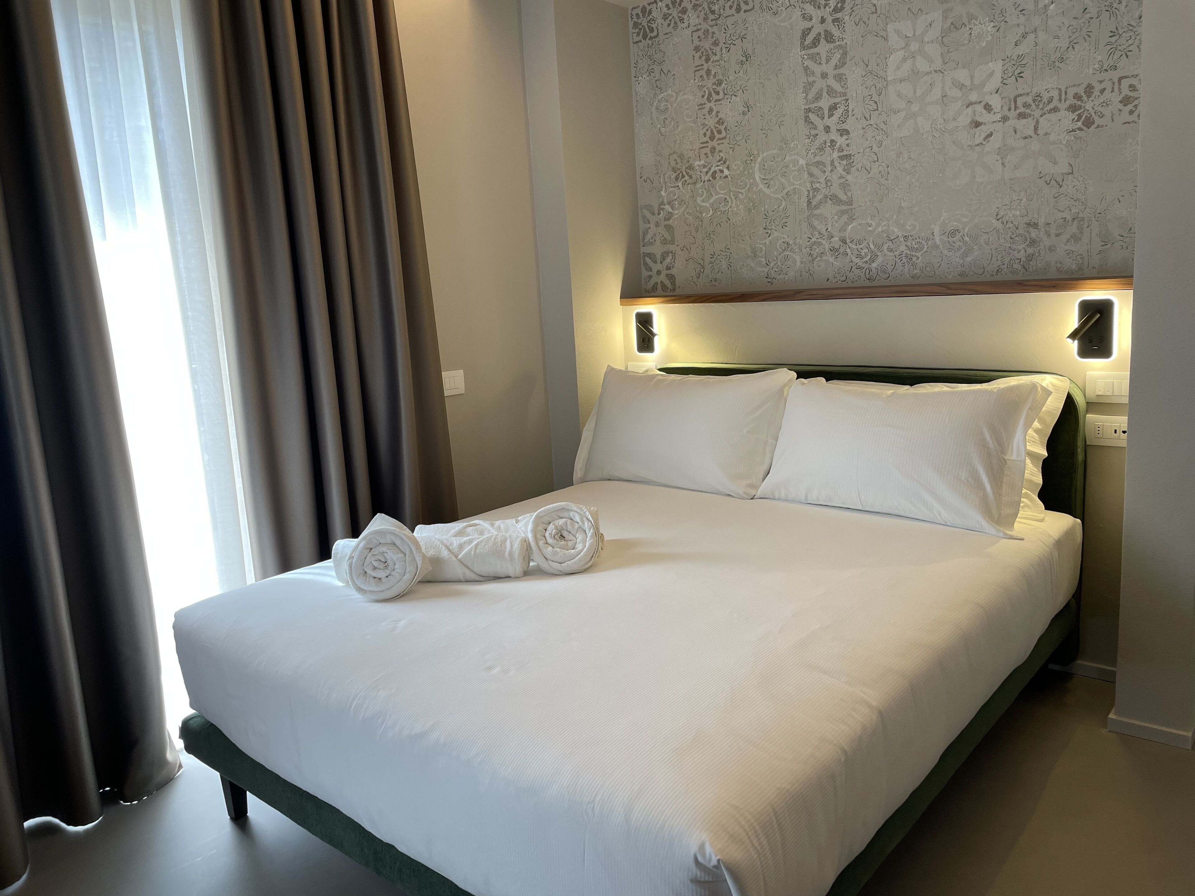 Un letto rifatto con lenzuola bianche e asciugamani arrotolati, una testiera con luci a parete e una parete decorata con un motivo.