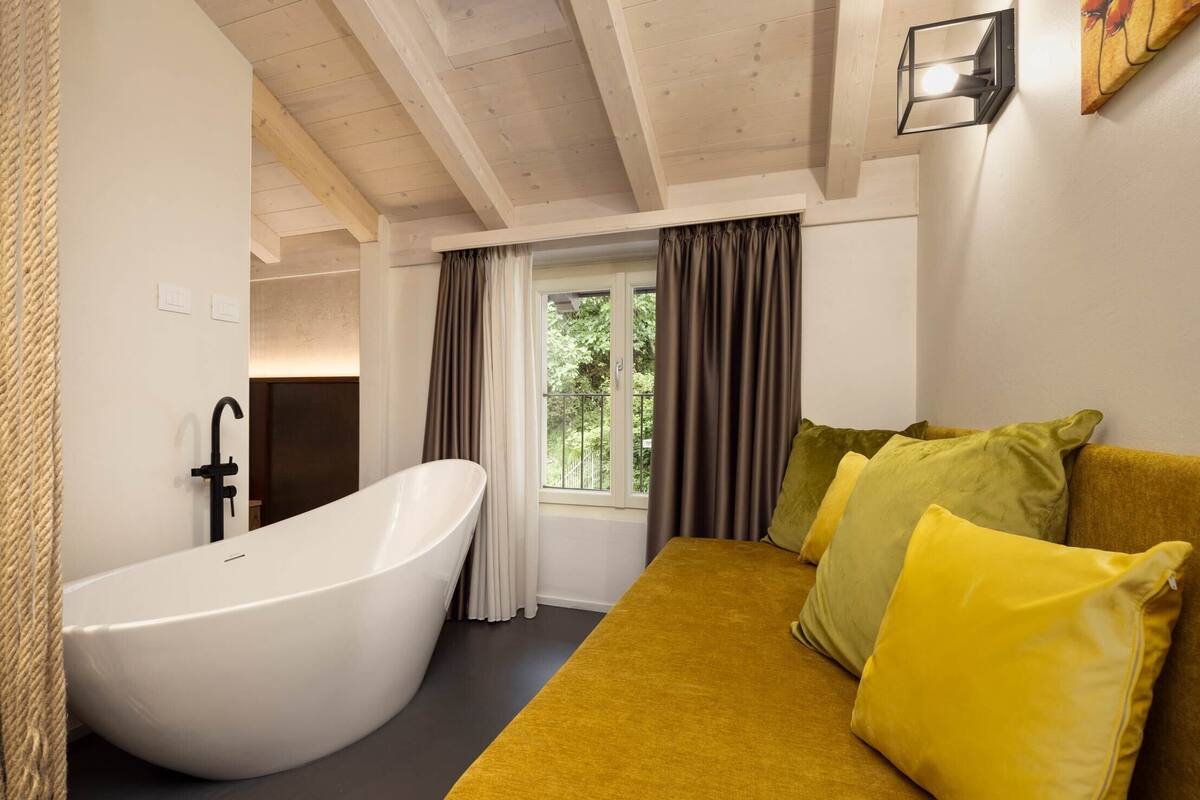 Ein Badezimmer mit einer freistehenden Badewanne, einem Fenster mit Vorhängen und einem senffarbenen Sofa mit gelben Kissen.