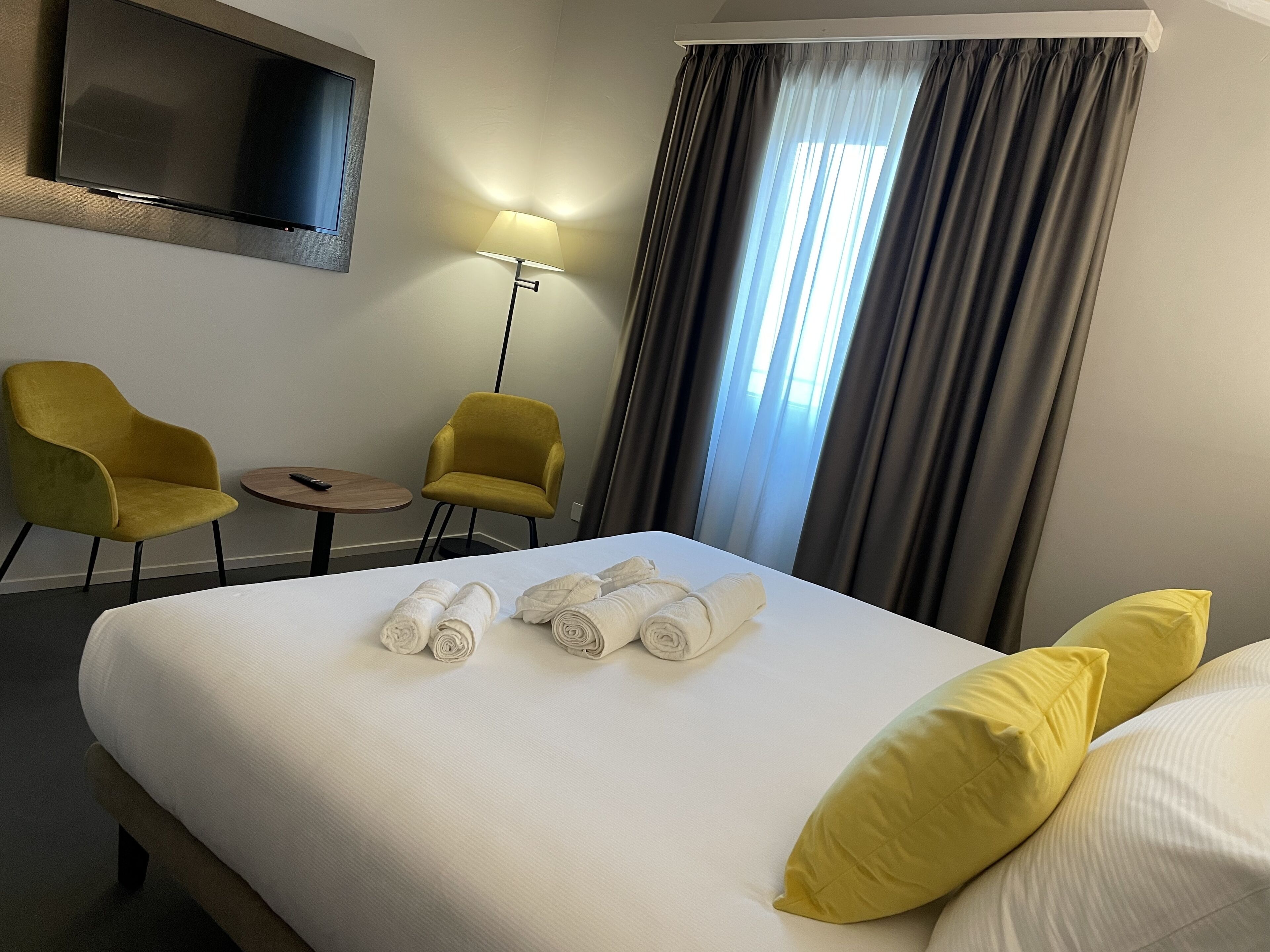 Una habitació d'hotel amb un llit, dues butaques grogues, una taula petita, una làmpada i un televisor fixat a la paret.
