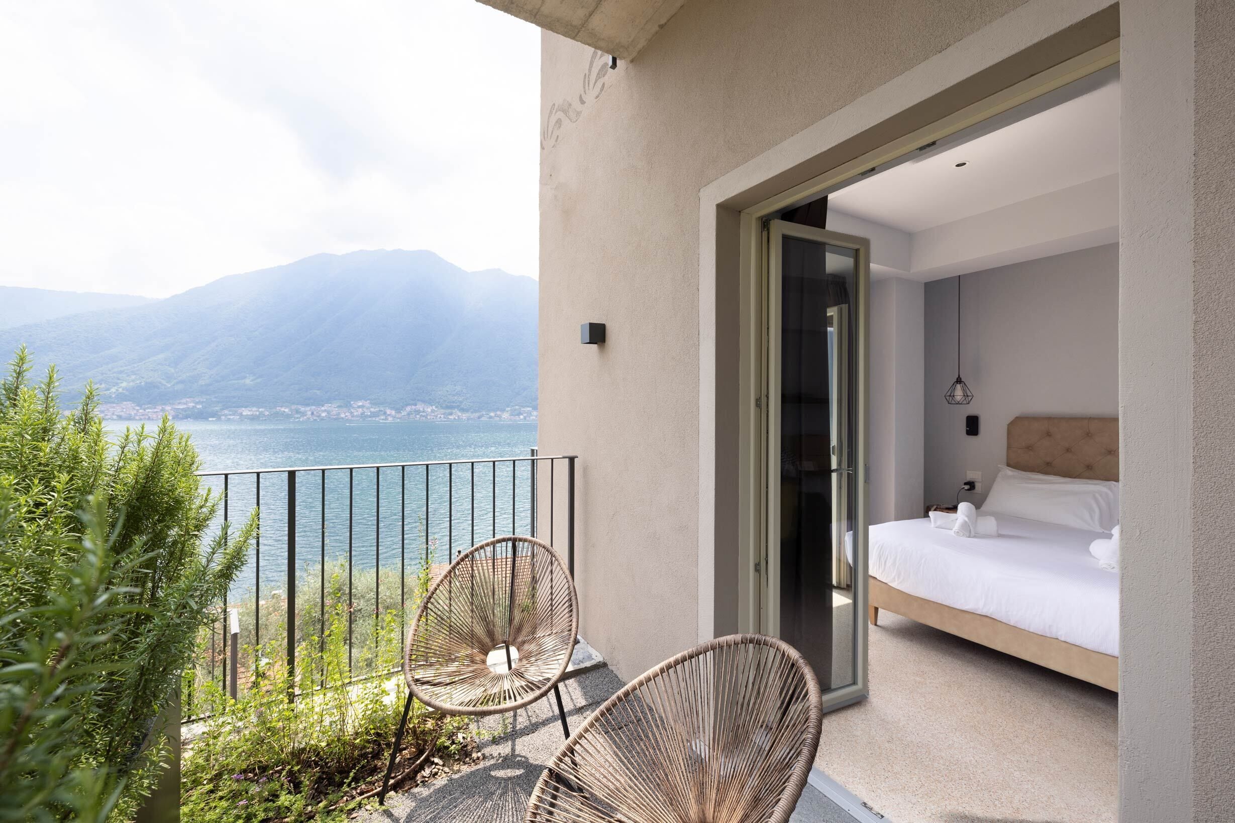 A balcony with wicker chairs, a bed with white linens, and a view of a lake and mountains.