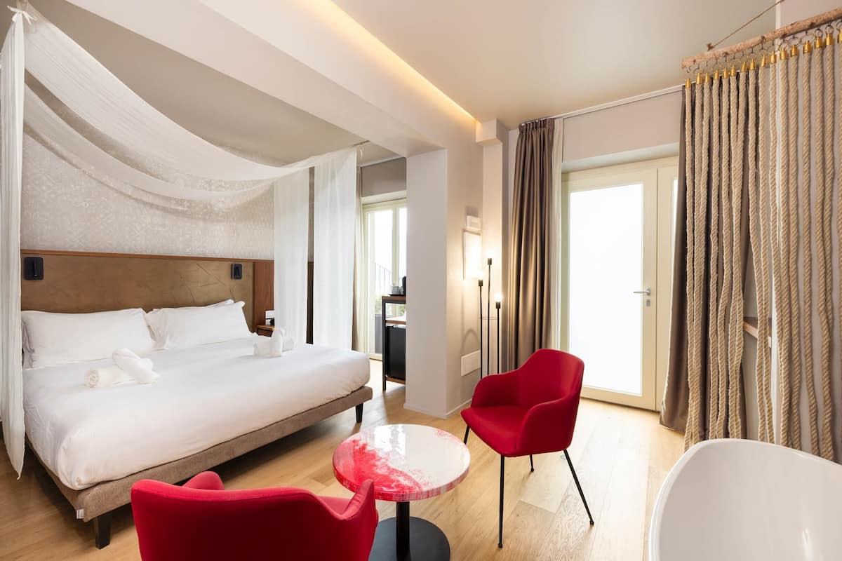 Ein Hotelzimmer mit einem großen Bett, einem roten Sessel, einem kleinen runden Tisch und Blick hinaus durch eine Tür.