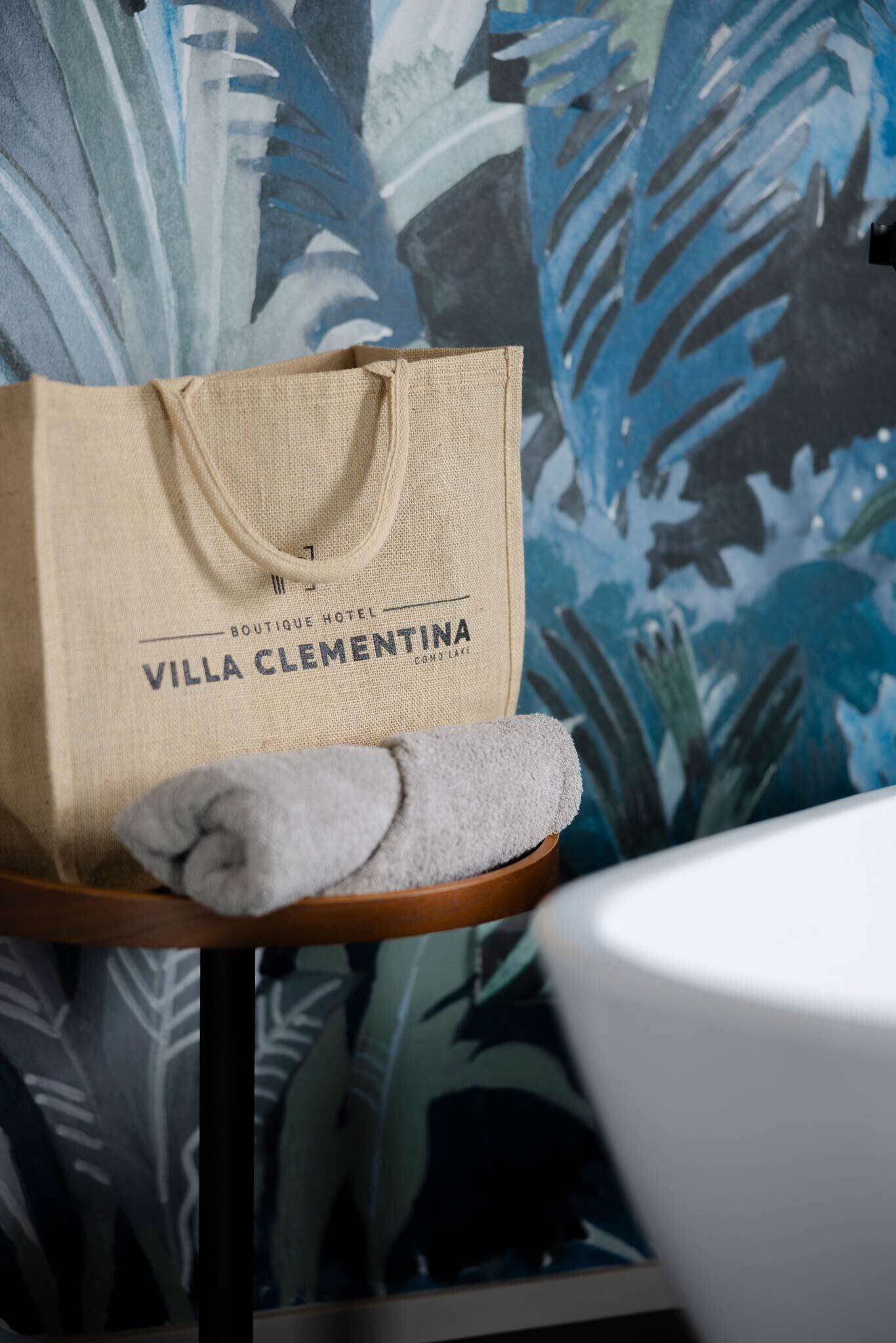 Una bolsa color beige con el texto "BOUTIQUE HOTEL VILLA CLEMENTINA" sobre una superficie de madera, una toalla gris y una bañera blanca frente a un fondo de papel tapiz floral.