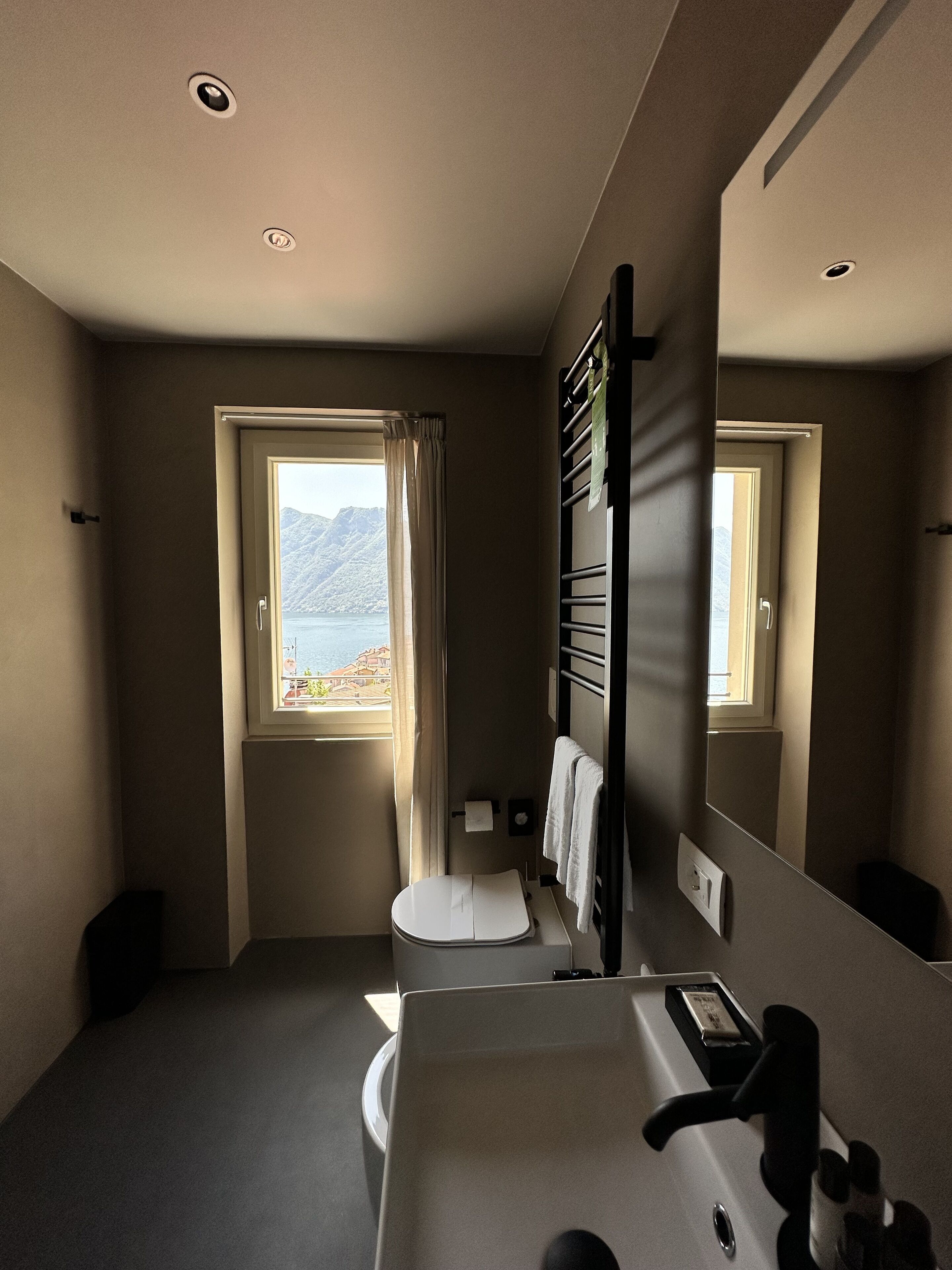 Un bagno moderno con il water, un lavandino e un grande specchio. Una finestra offre vista su montagne e spiaggia.