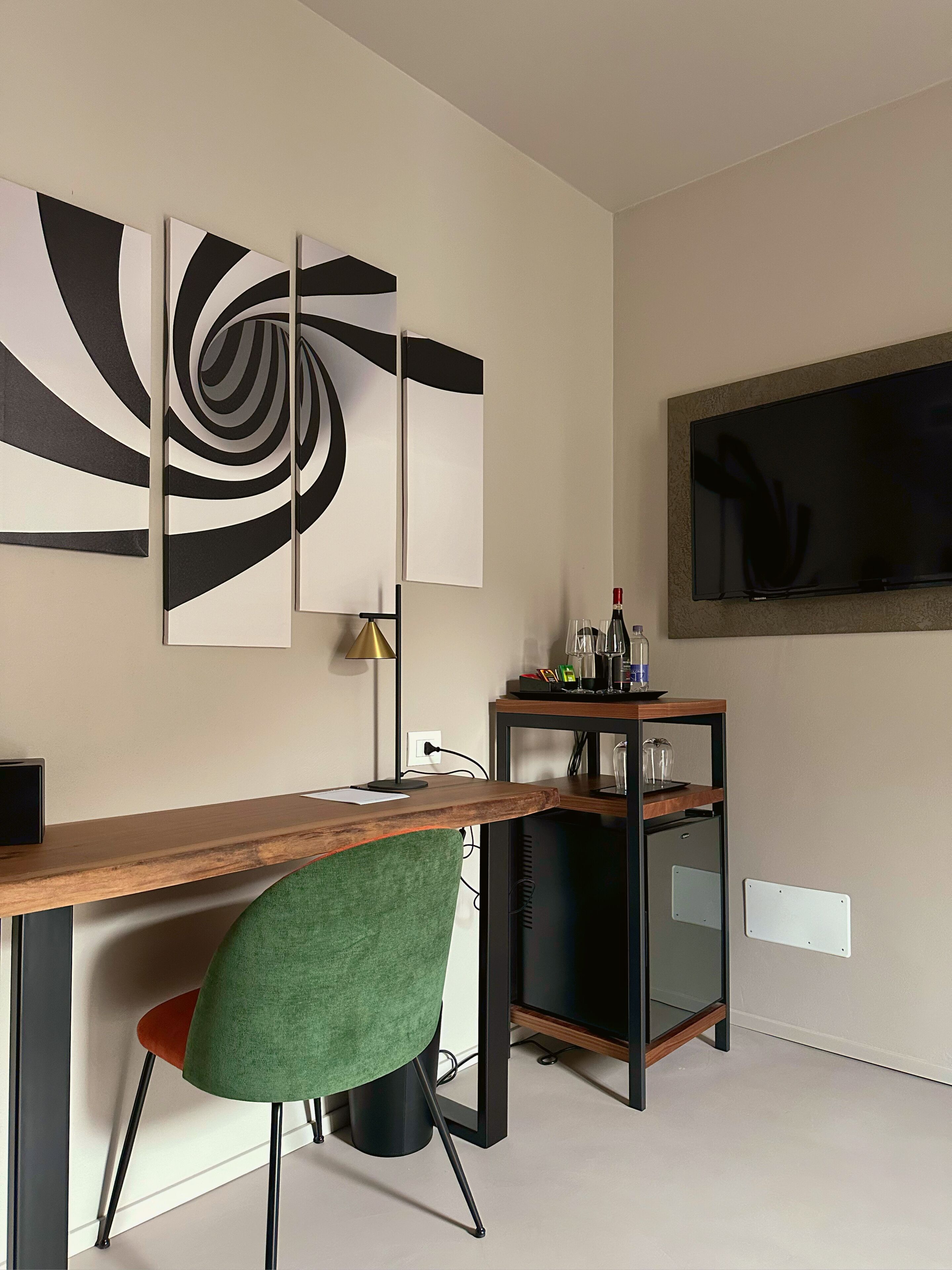 Un ufficio moderno con scrivania in legno, una sedia verde, un quadro astratto in bianco e nero, un televisore montato a parete e una piccola mensola con bicchieri e una bottiglia.