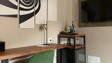 Un ufficio moderno con scrivania in legno, una sedia verde, un quadro astratto in bianco e nero, un televisore montato a parete e una piccola mensola con bicchieri e una bottiglia.