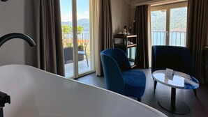 A modern bathroom with a freestanding bathtub, a seating area with two chairs, a small round table, and a balcony with a view of the mountains. - Villa Clementina Boutique Hotel (Colonno)