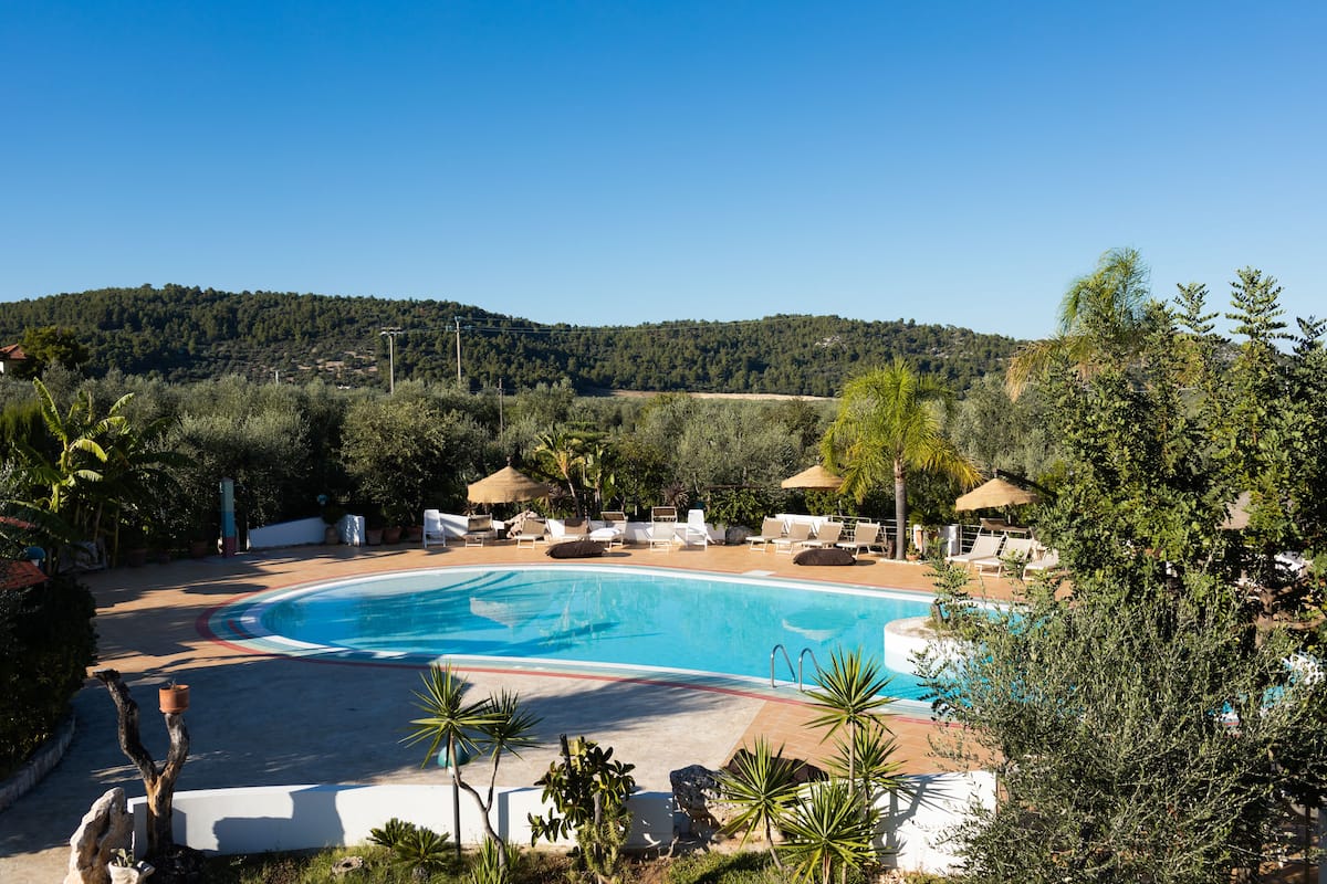 Piscina com espreguiçadeiras e guarda-sóis, cercada por vegetação e colinas ao fundo.
