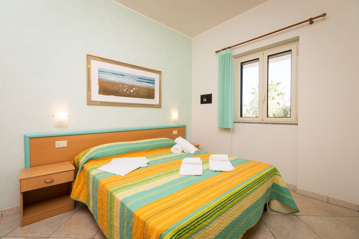 Quarto com cama, mesa de cabeceira, luminária na parede, janela com cortinas e um quadro na parede.