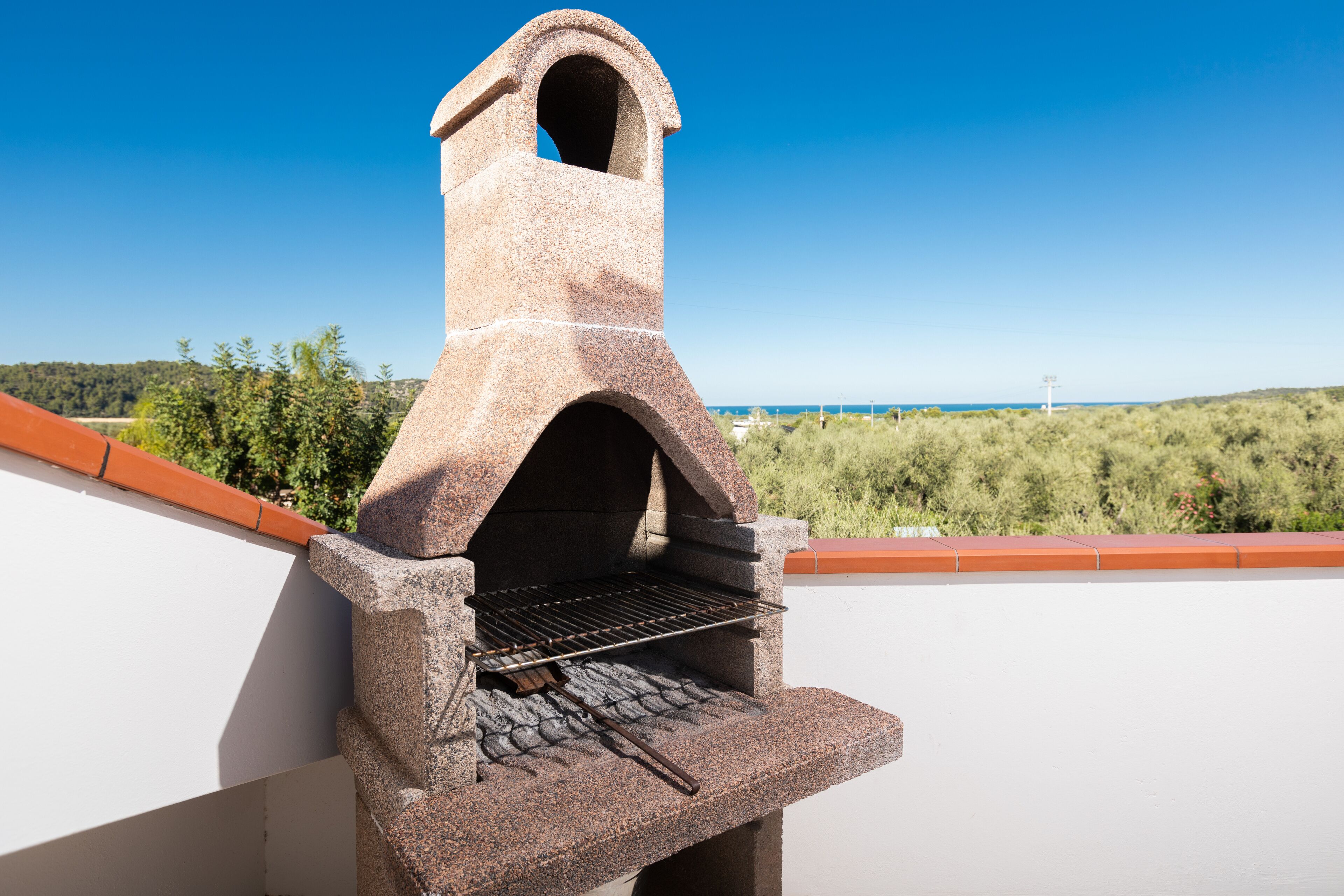 A stone-built outdoor grill with a chimney, set against a clear blue sky and a view of a forested landscape.