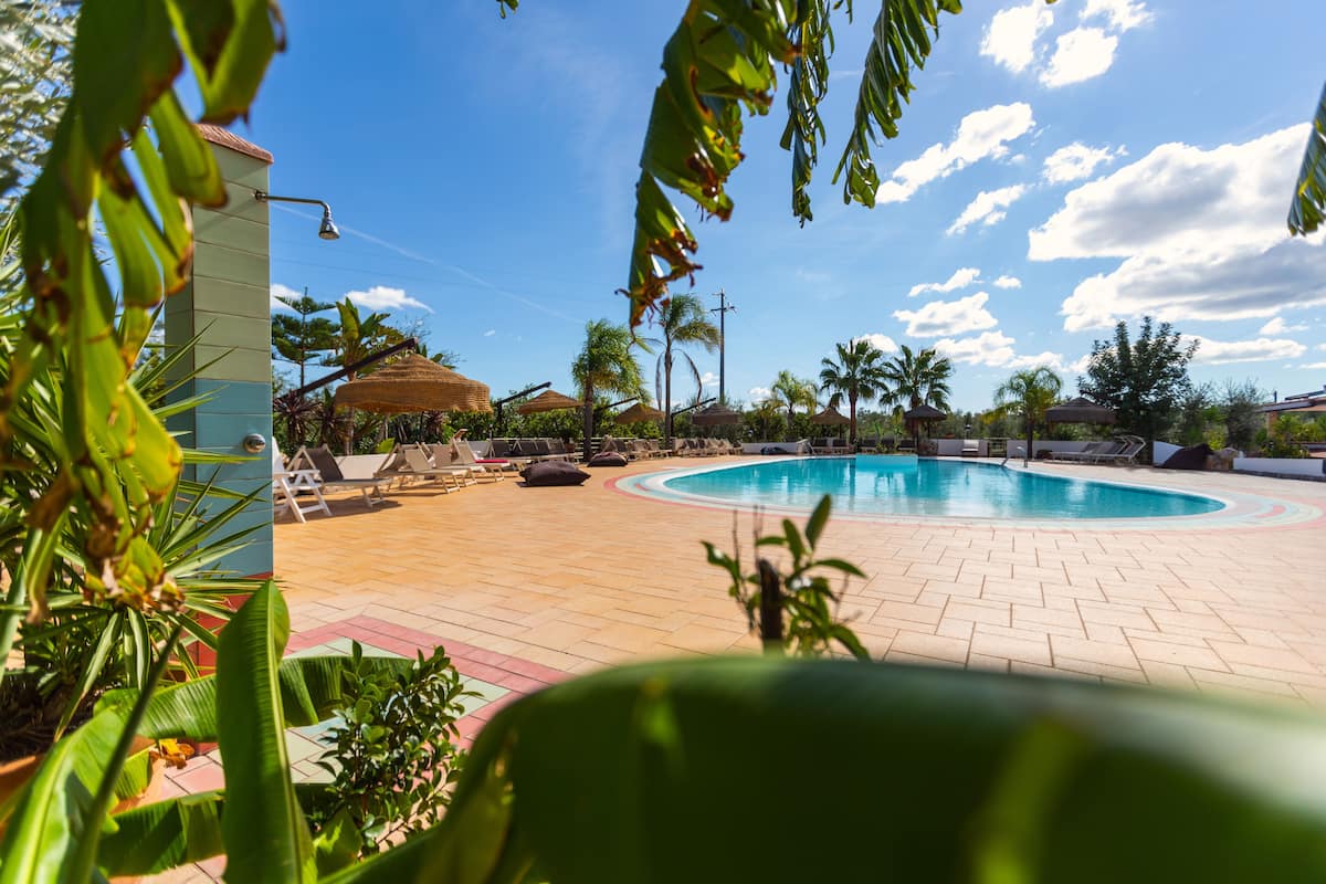 Piscina com espreguiçadeiras e palmeiras, rodeada por edifícios e céu claro.