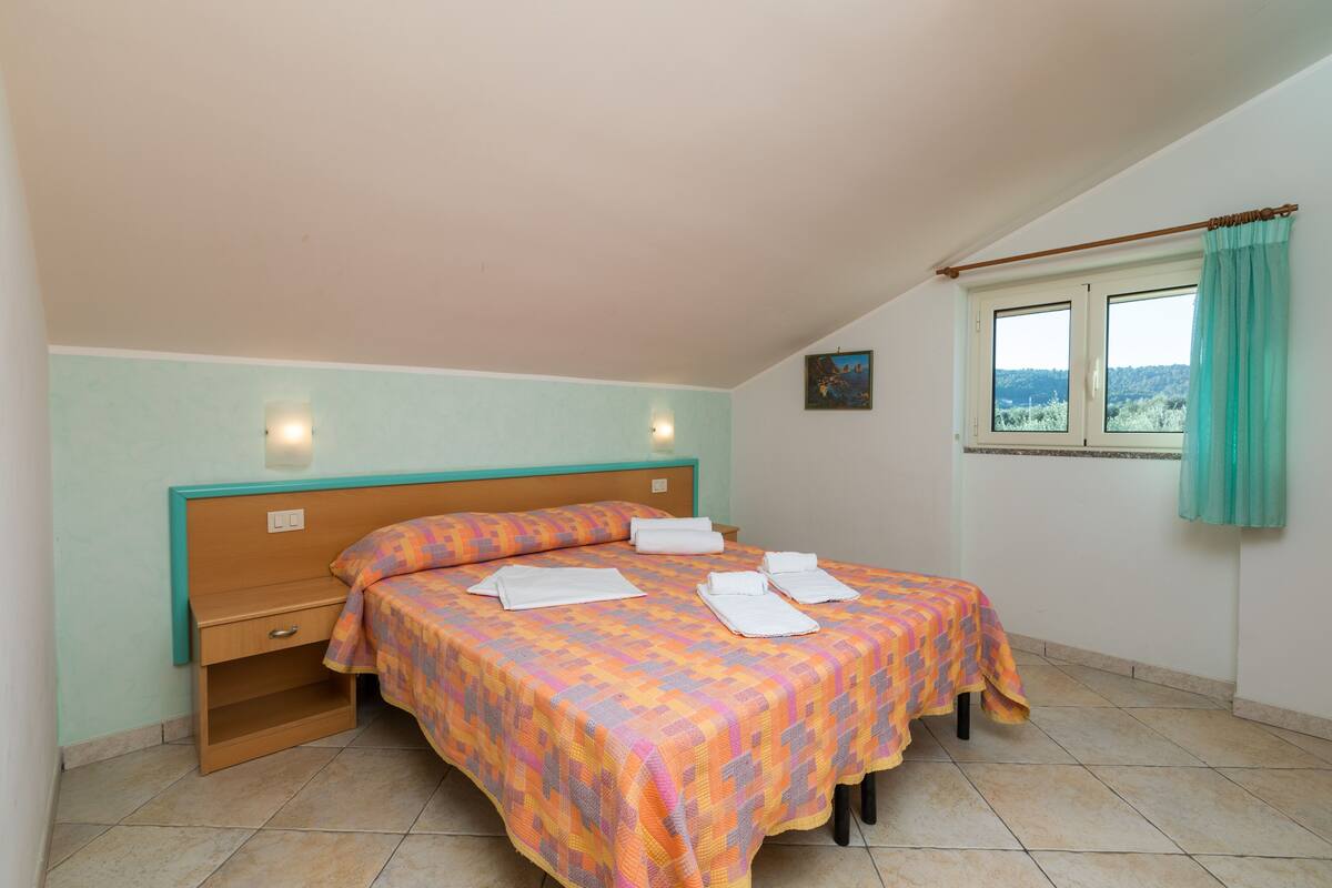 Quarto com cama, mesa de cabeceira, janela com cortinas e vista para o exterior.