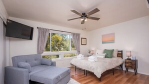 A bedroom with a bed, a sofa, a TV, a ceiling fan, and a window with curtains.