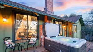 A hot tub on a wooden deck with a covered patio area, a brick chimney, and a view of the interior through large windows.
