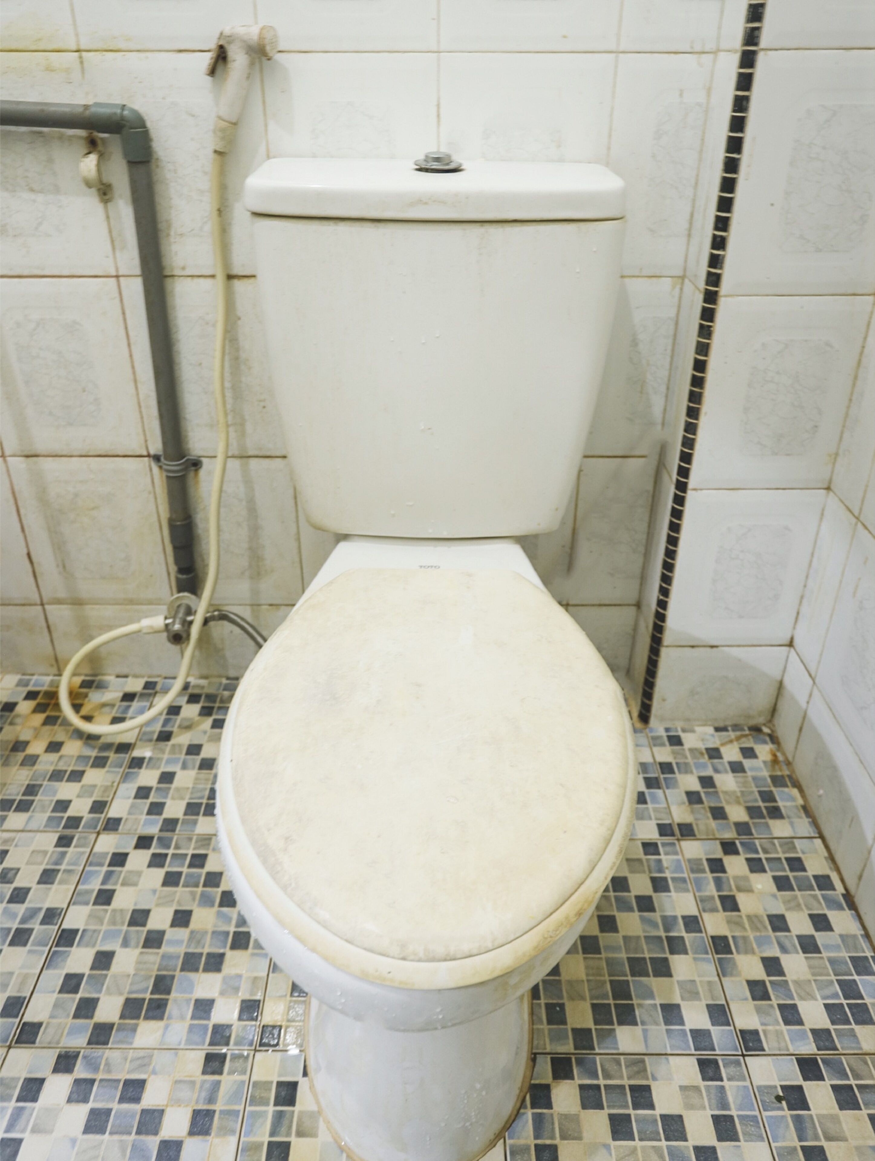 A toilet with a white seat and lid, a visible pipe, and a tiled floor with a checkered pattern.