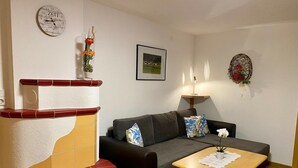 A living room with a sofa, a small wooden table, a wall clock, a framed picture, and a decorative wall piece. - Ihr Zuhause im Urlaub - Familiär und Komfortabel (Niederthai)