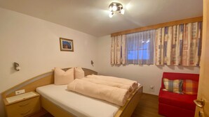 A bedroom with a wooden bed, a nightstand, a red chair, and a window with curtains. - Ihr Zuhause im Urlaub - Familiär und Komfortabel (Niederthai)