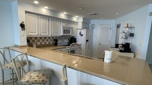 A modern kitchen with white cabinets, a granite countertop, a built-in oven, a microwave, and a coffee maker. There are two bar stools with patterned cushions.
