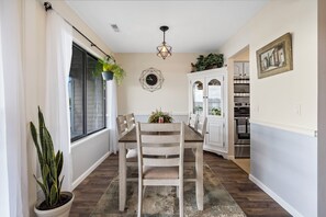 Un coin repas comprenant une table en bois, des chaises, un luminaire suspendu, une horloge et un meuble avec des portes en verre.