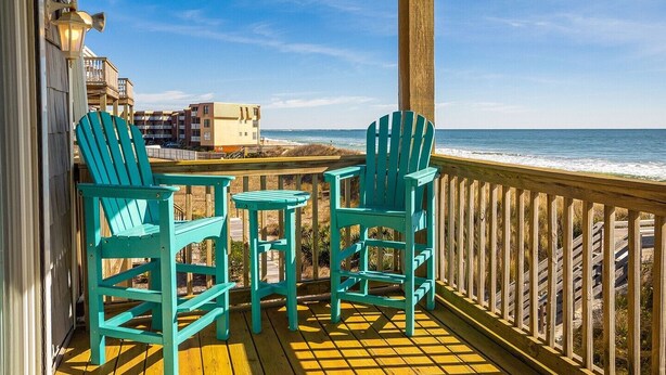 Una terraza de madera con dos sillas Adirondack turquesa y una mesita, con vista a una playa y edificios al fondo.