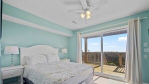 Un dormitorio con cama, mesitas de noche, ventilador de techo y un amplio ventanal corredera que da a un balcón.