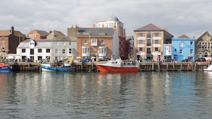 A waterfront with boats docked, a variety of buildings including a blue marine engineering office, and a row of shops and eateries along the pier. - Embassy (Weymouth)
