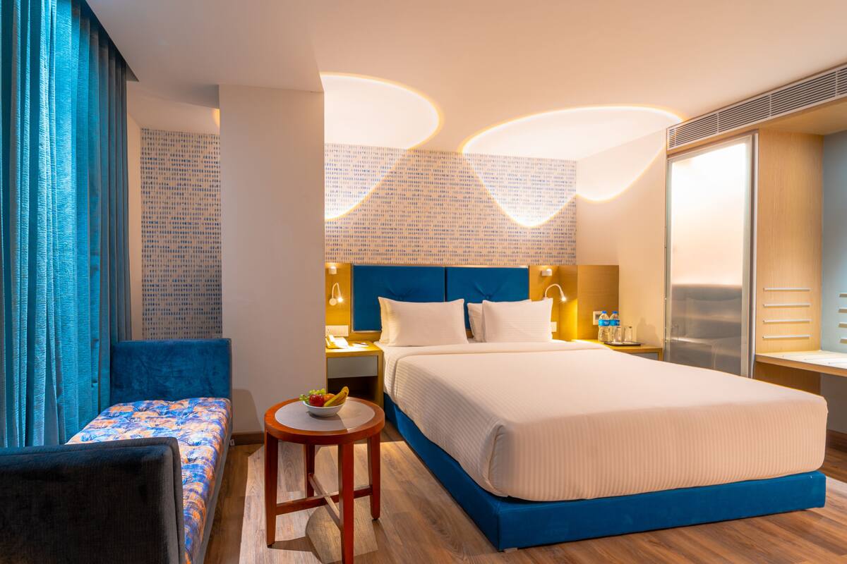 Una moderna camera d'hotel con un letto grande, un comodino con una lampada, un tavolino rotondo con della frutta e un divano blu.
