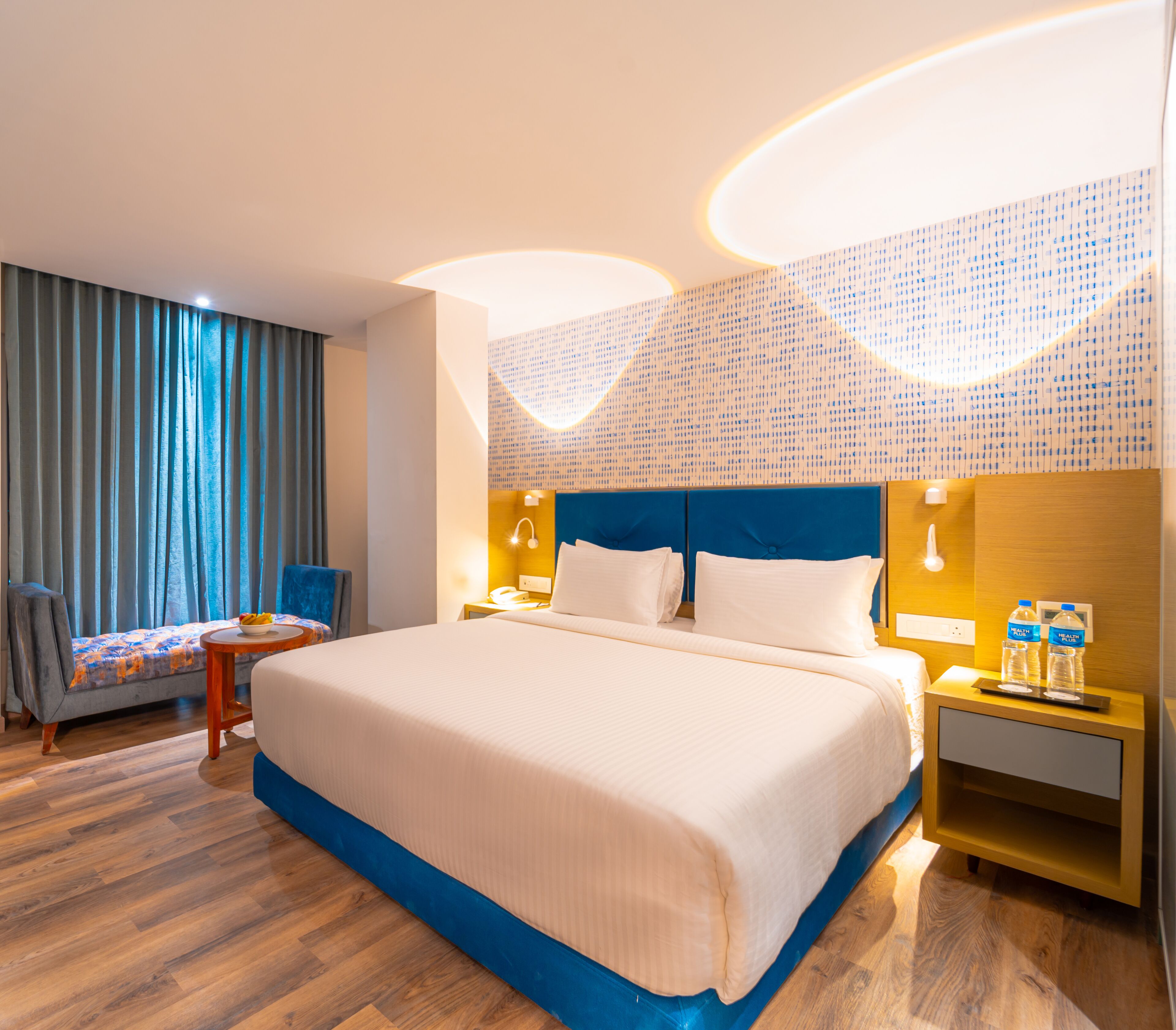A hotel room with a large bed, a chair, a small table, and a nightstand with water bottles.