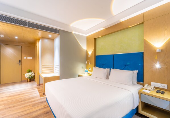 Una habitación de hotel moderna con una cama grande, suelo de madera, una pared azul como acento y una mesita de noche con teléfono y lámpara.