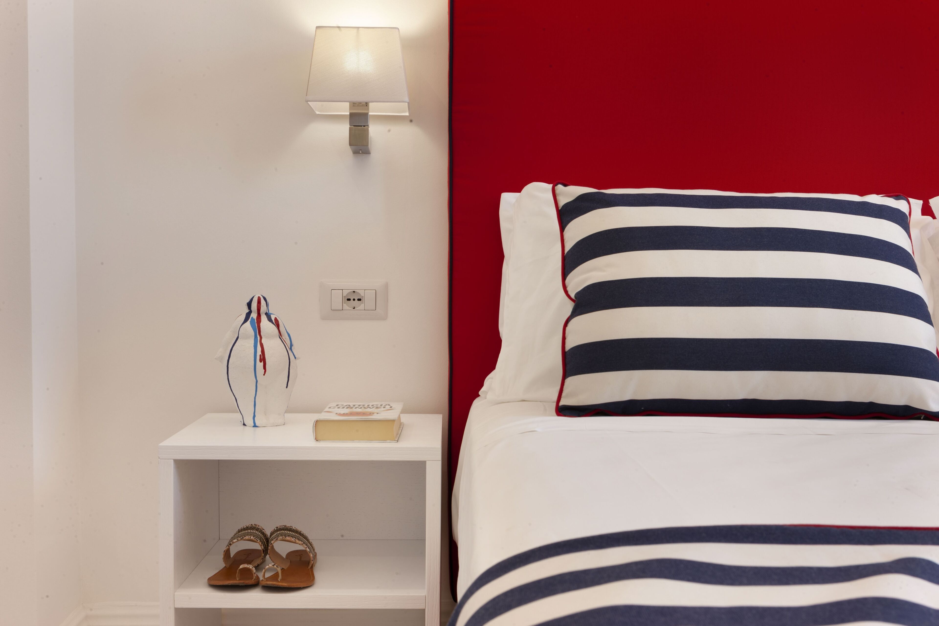 A neatly made bed with striped bedding, a bedside lamp, a small nightstand with a book and a pair of sandals, and a red accent wall.
