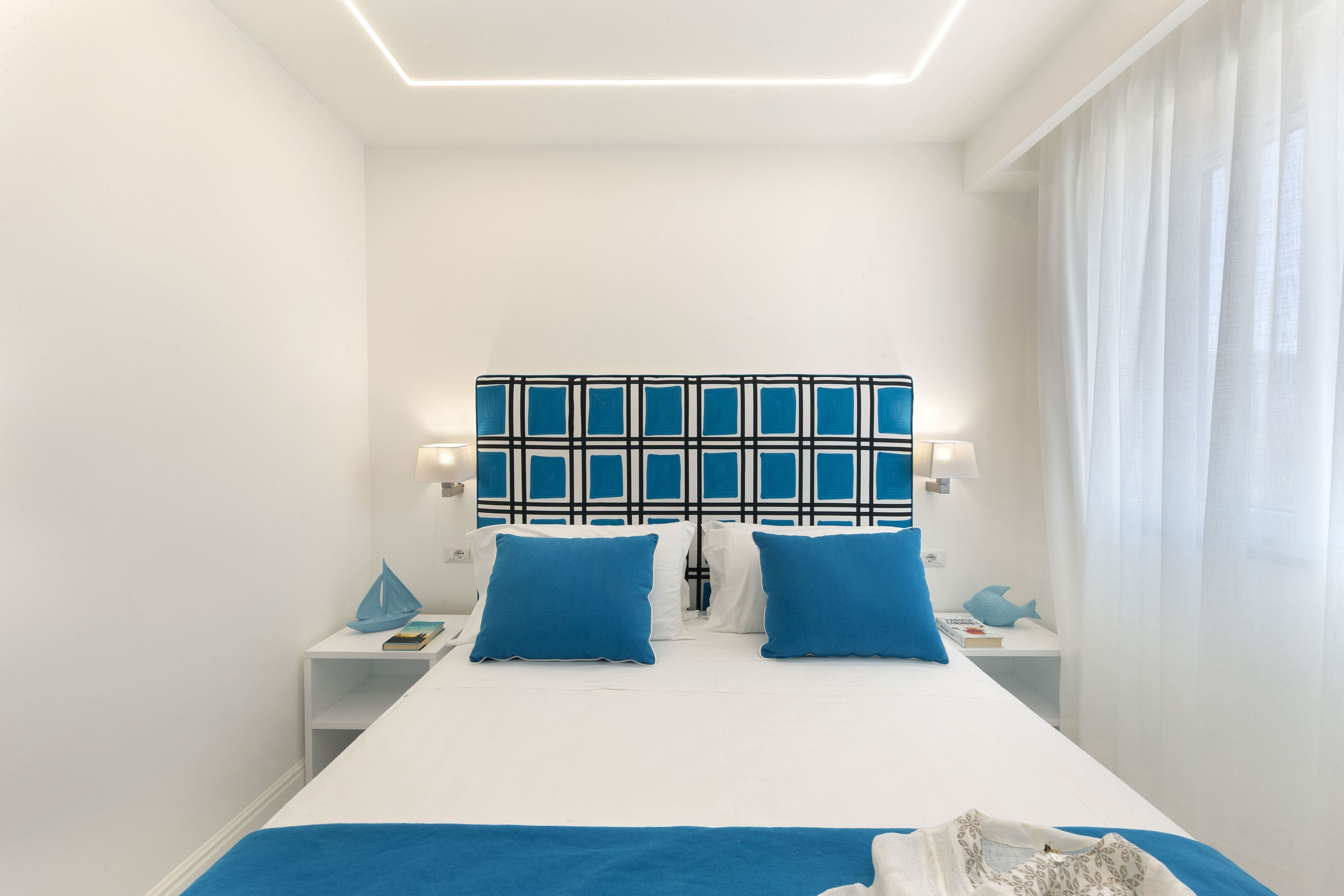 Una habitación de hotel moderna con una cama grande, dos almohadas azules, un cabecero estampado y una mesita de noche blanca con una lámpara.