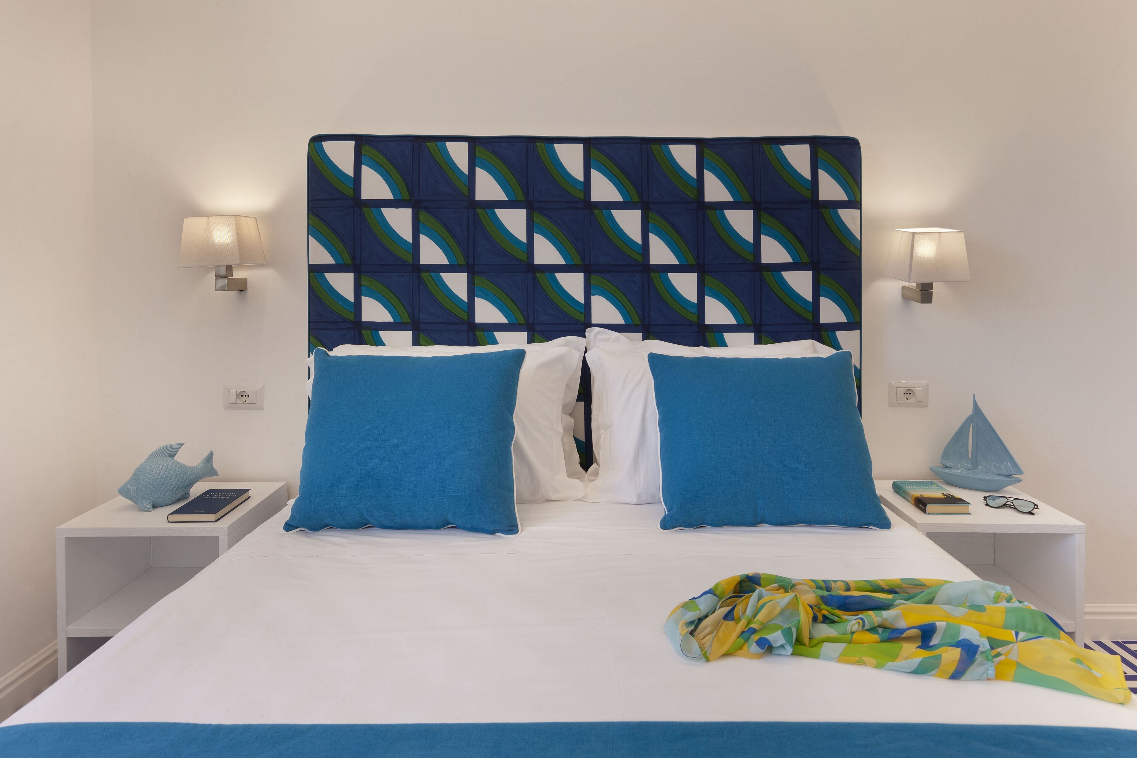 A neatly made bed with blue and white bedding, two bedside tables with lamps, a small shelf with books and a decorative item, and a colorful blanket on the bed.