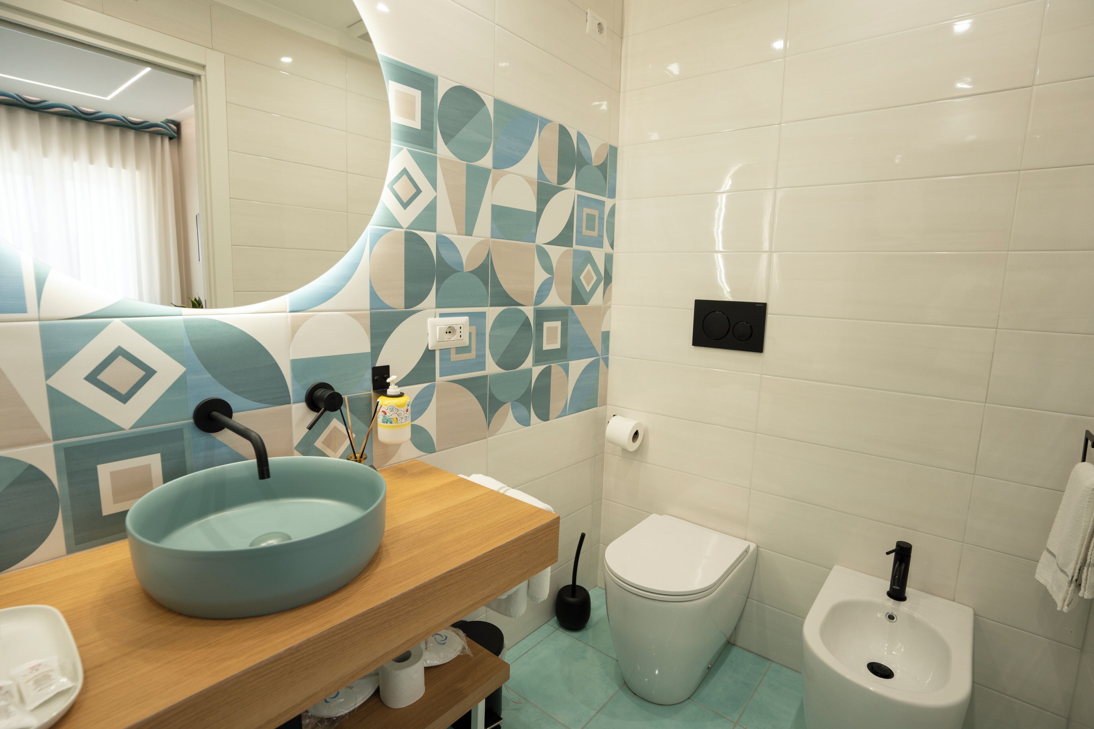 A bathroom with a sink, toilet, bidet, and a mirror with a geometric tile backsplash.