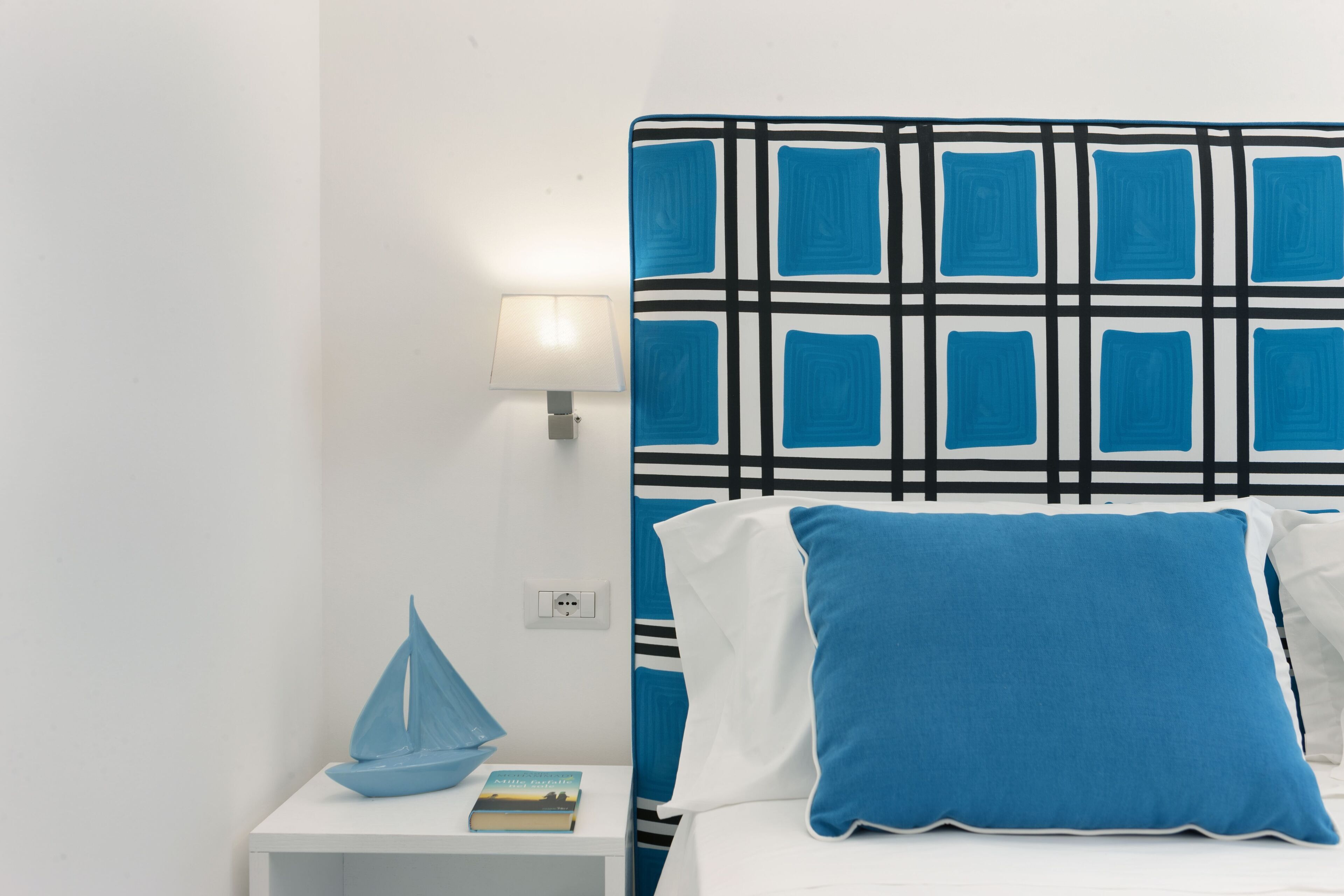 A bedroom with a white bed, blue pillows, a blue sailboat decoration, and a wall-mounted lamp.