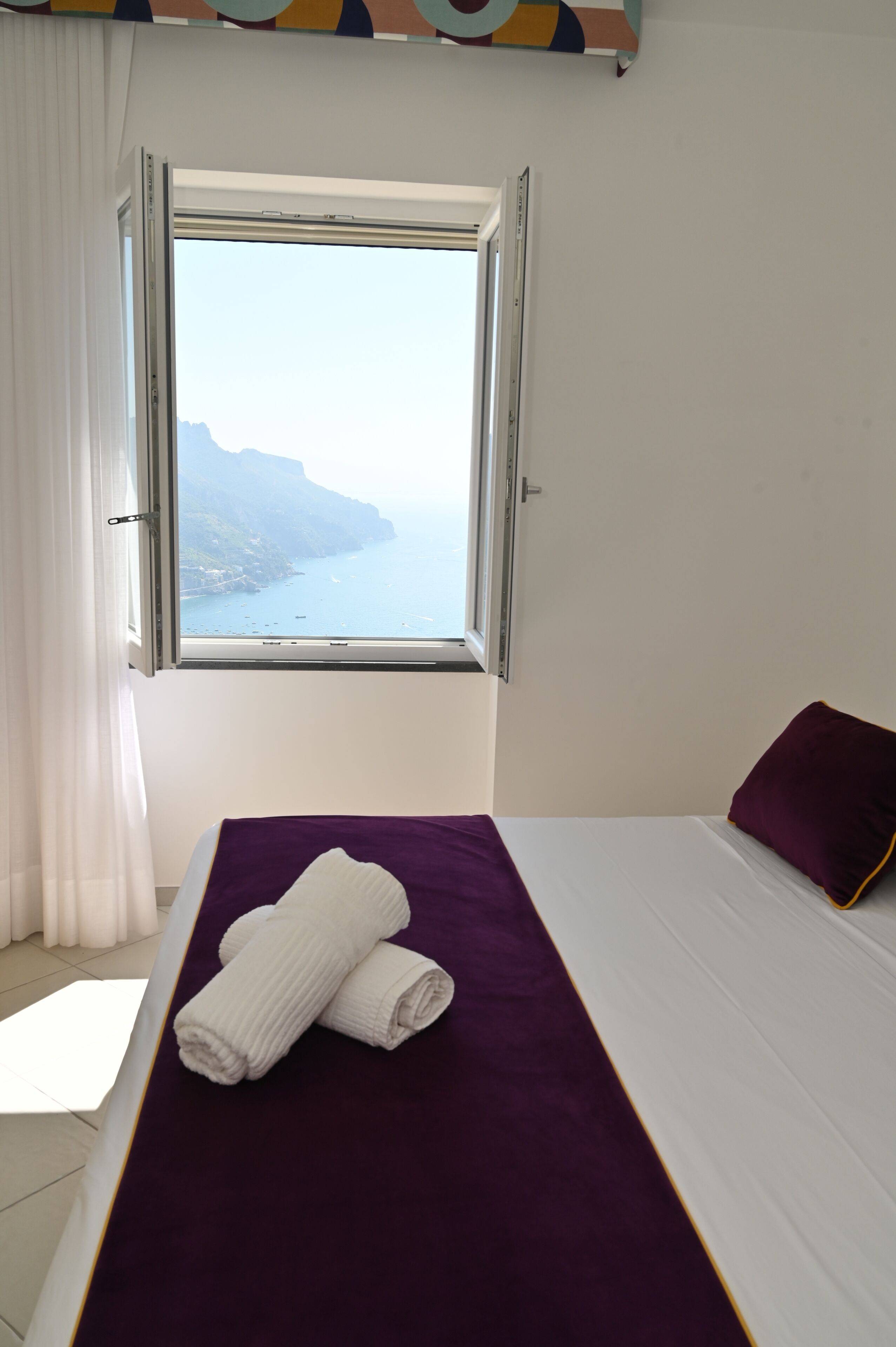 Ett hotellrum med en säng, ett fönster med utsikt över havet och klipporna, och ett lila sängöverkast.