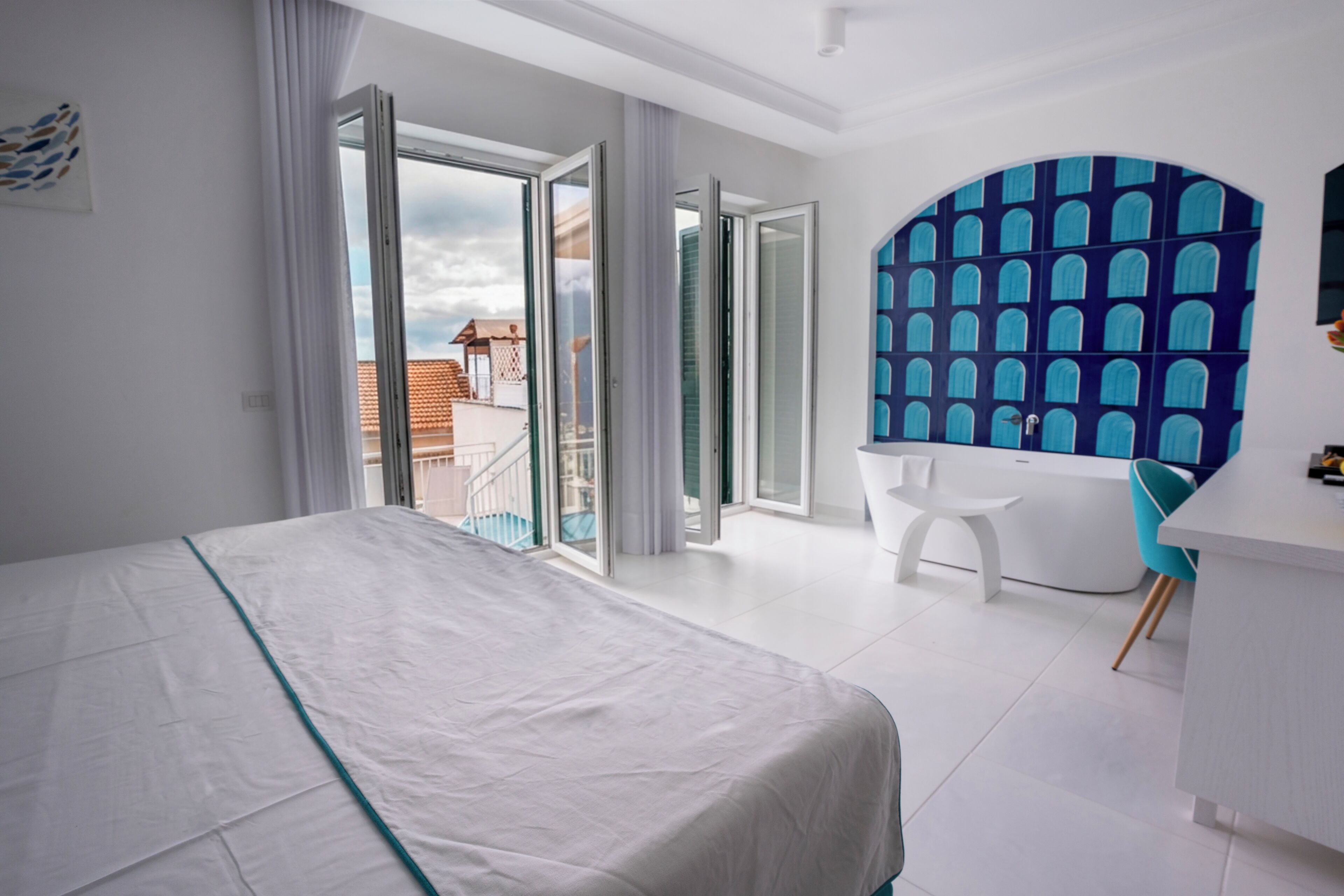 A modern hotel room with a large bed, a bathroom with a blue tiled wall, and a balcony with a view of buildings.