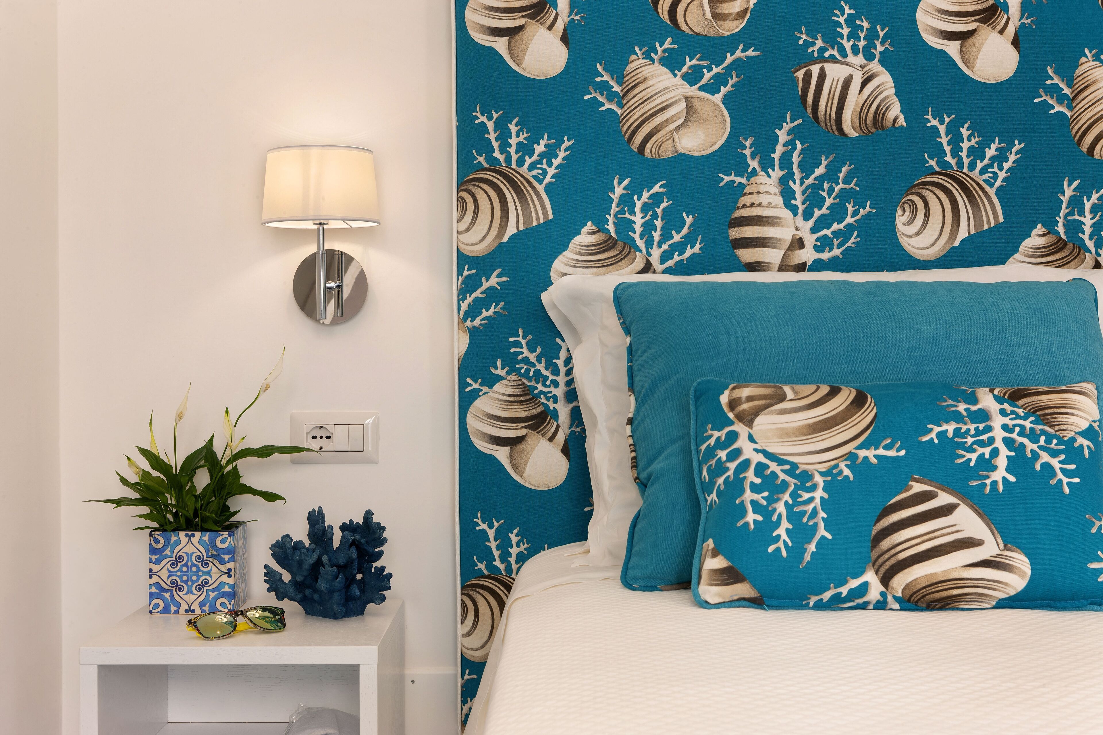 Camera da letto con carta da parati a motivi blu e bianchi a forma di conchiglia, un letto bianco con una coperta verde acqua, una lampada da comodino, una piccola pianta in vaso decorativo e una decorazione corallo sul comodino.