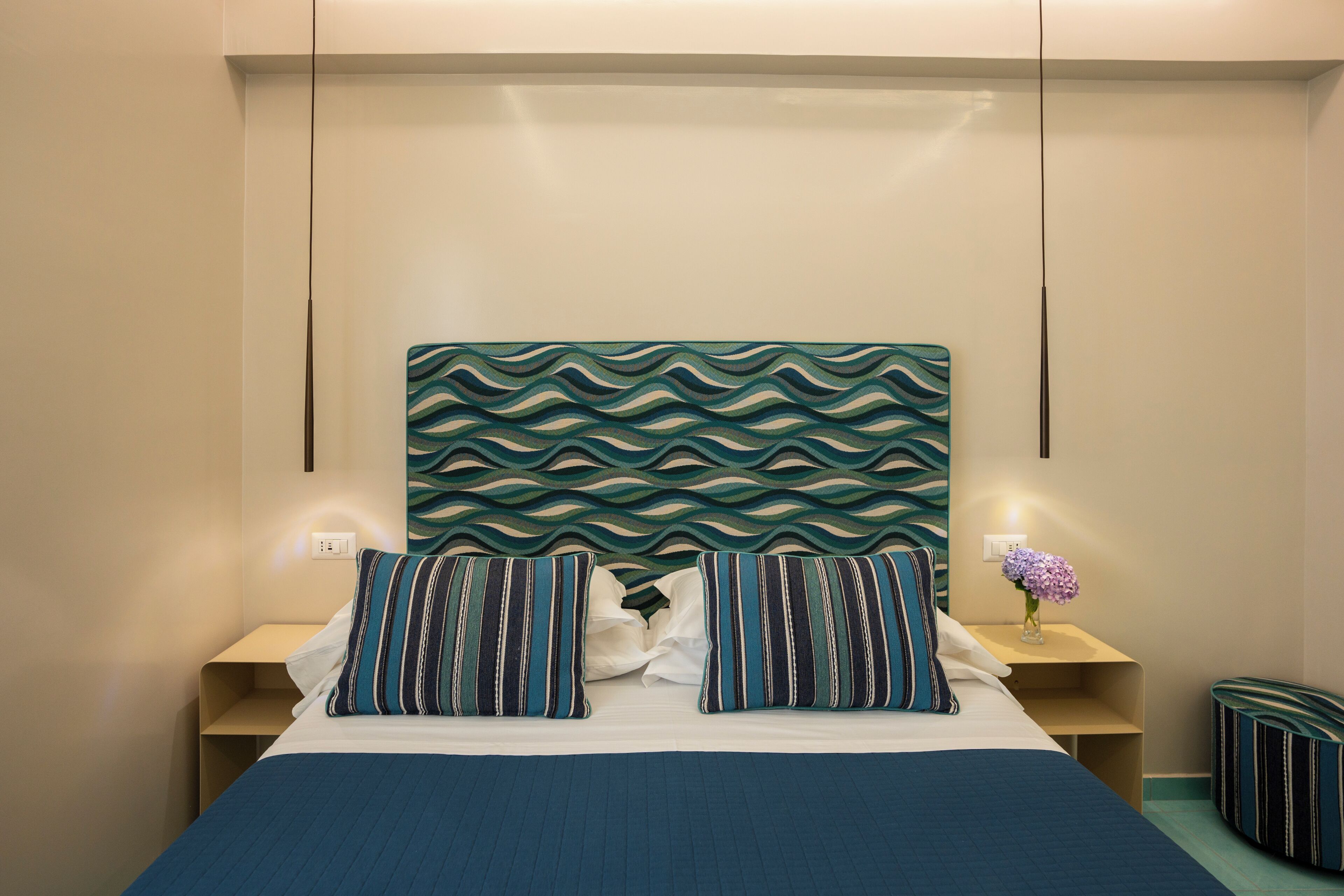 A neatly made bed with striped pillows, a headboard with a wavy pattern, bedside tables with lamps, and a small vase with flowers.