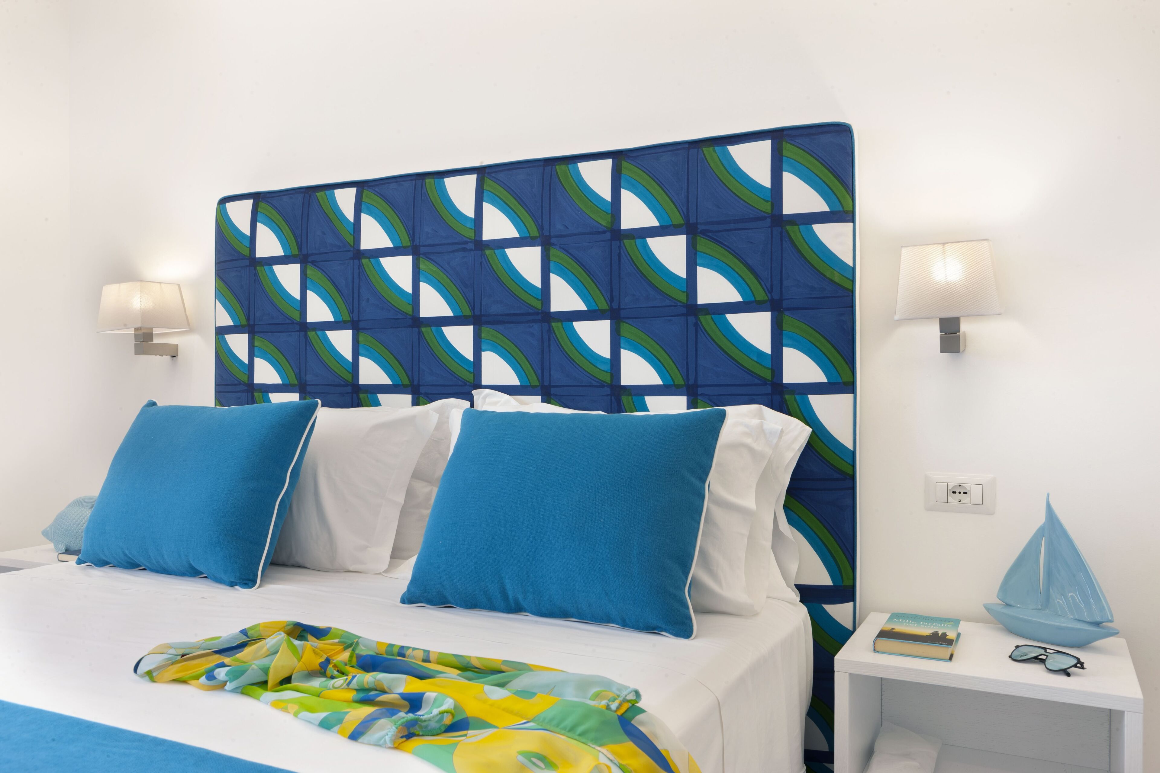 A bed with blue and white bedding, a colorful throw blanket, and a nightstand with a book and a blue sailboat model.