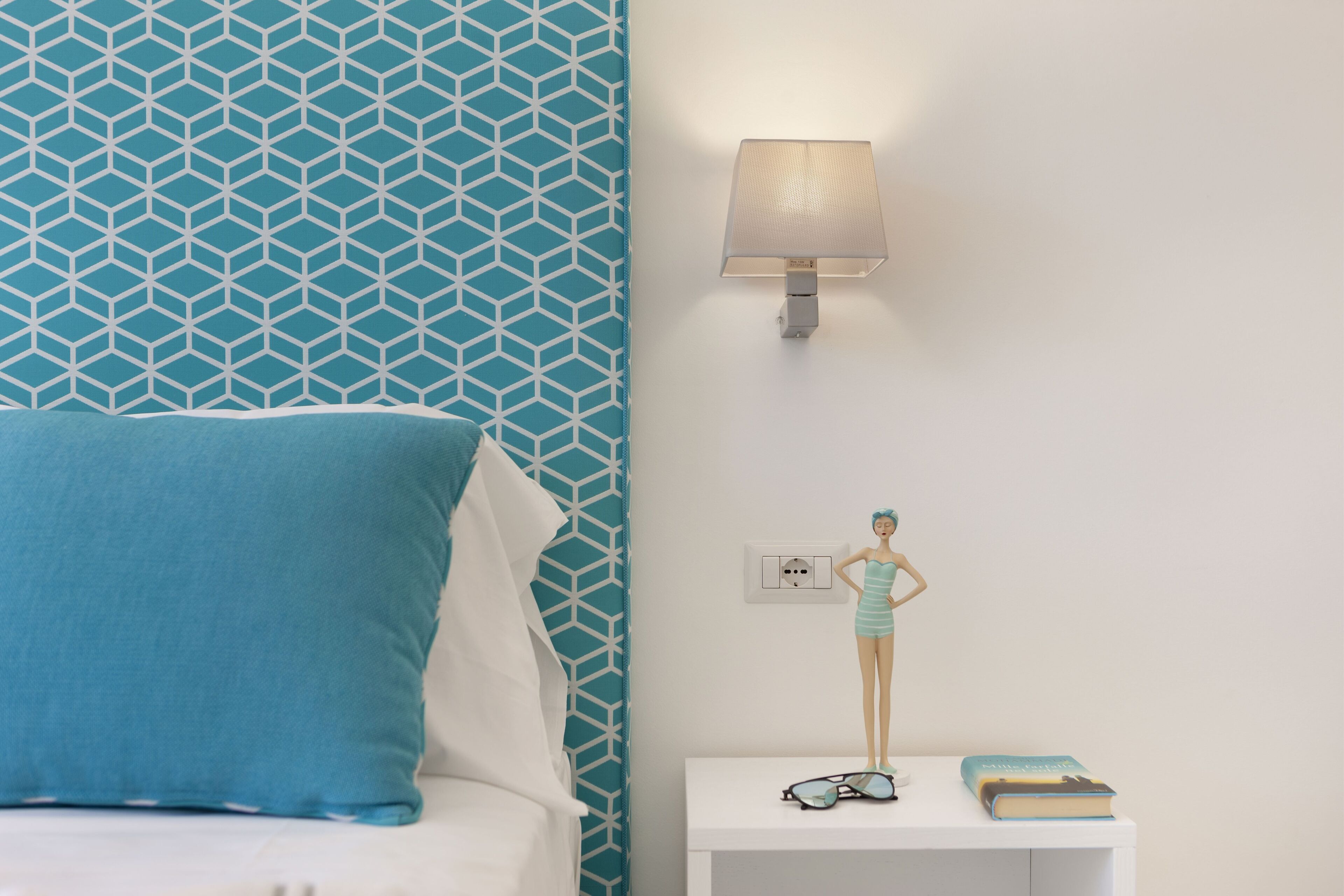 Une chambre avec un mur à motifs, une lampe de chevet, une table de chevet avec une poupée, et un lit avec un oreiller bleu.