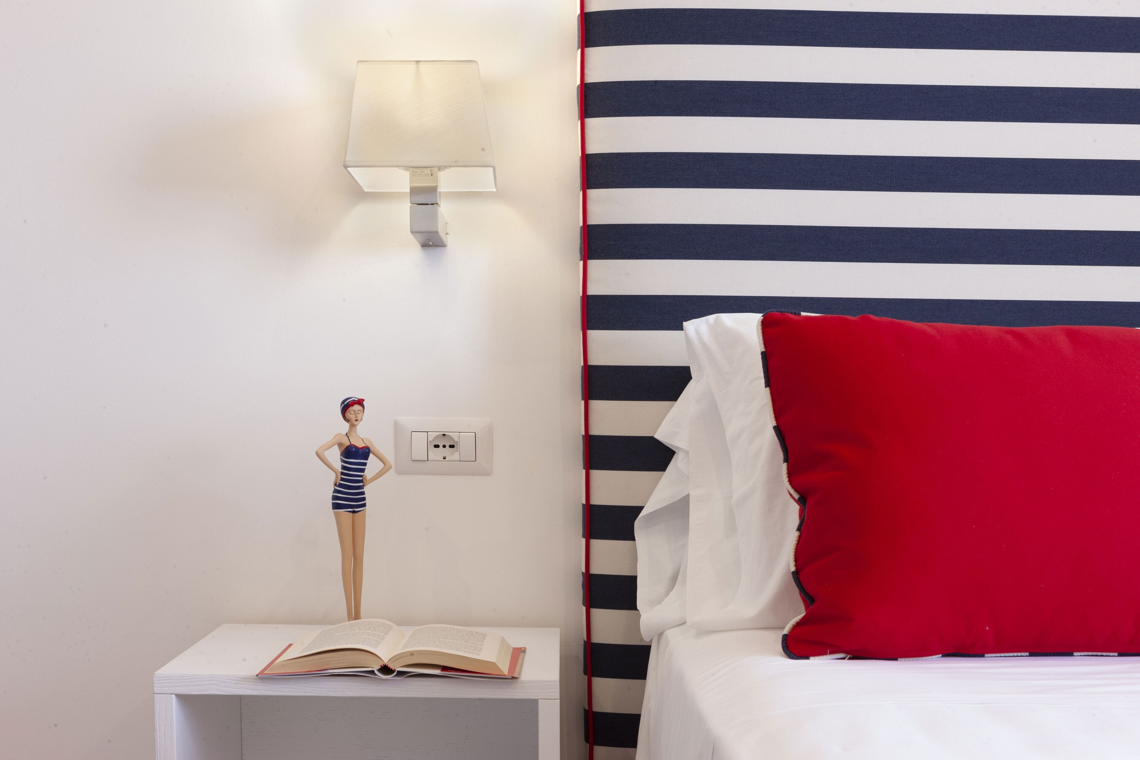 Una habitación con cabecera a rayas, almohada roja, mesita de noche con lámpara y un libro.