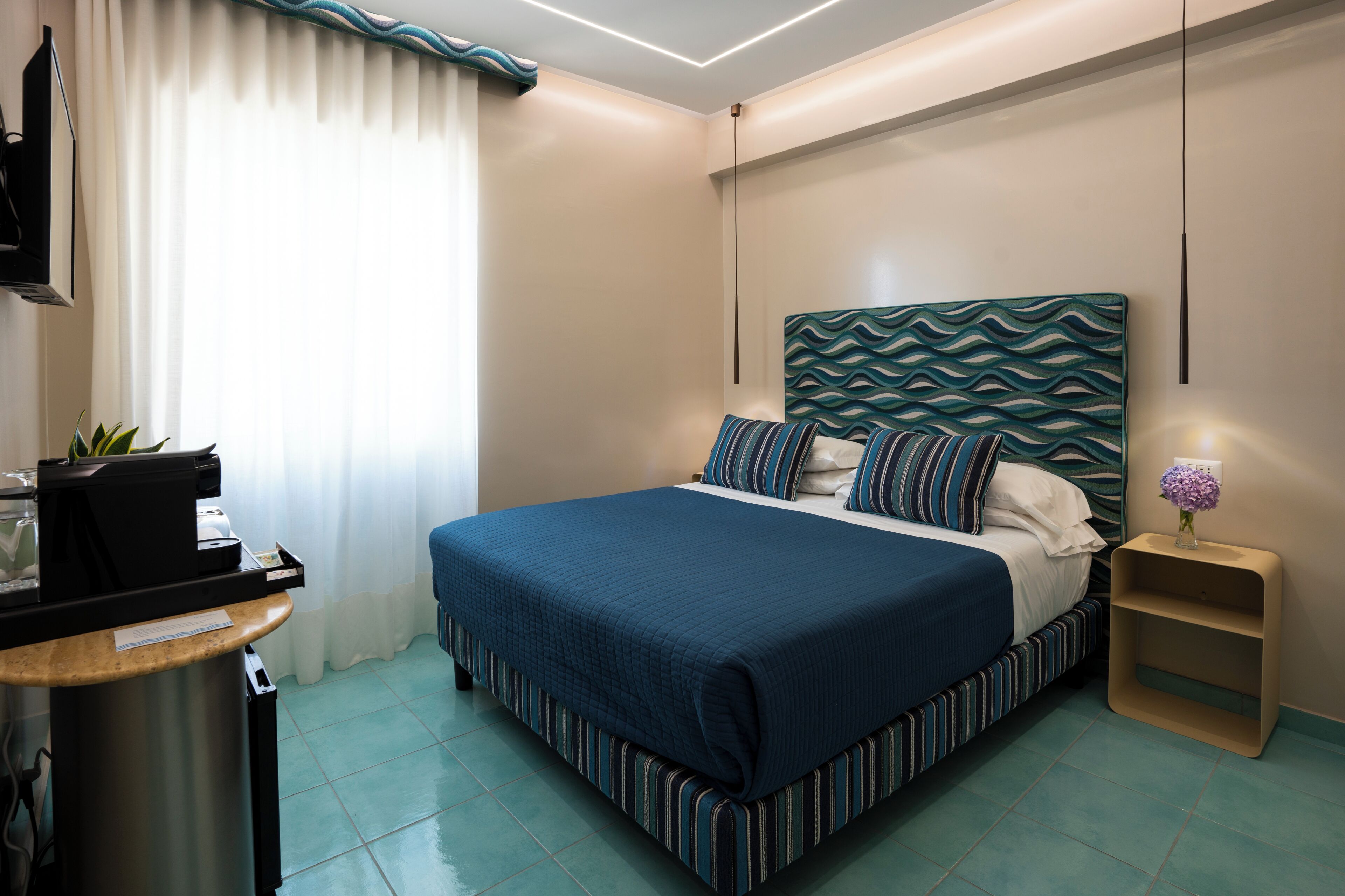 A hotel room with a bed, a desk, a chair, a small table, a lamp, and a wall-mounted TV.