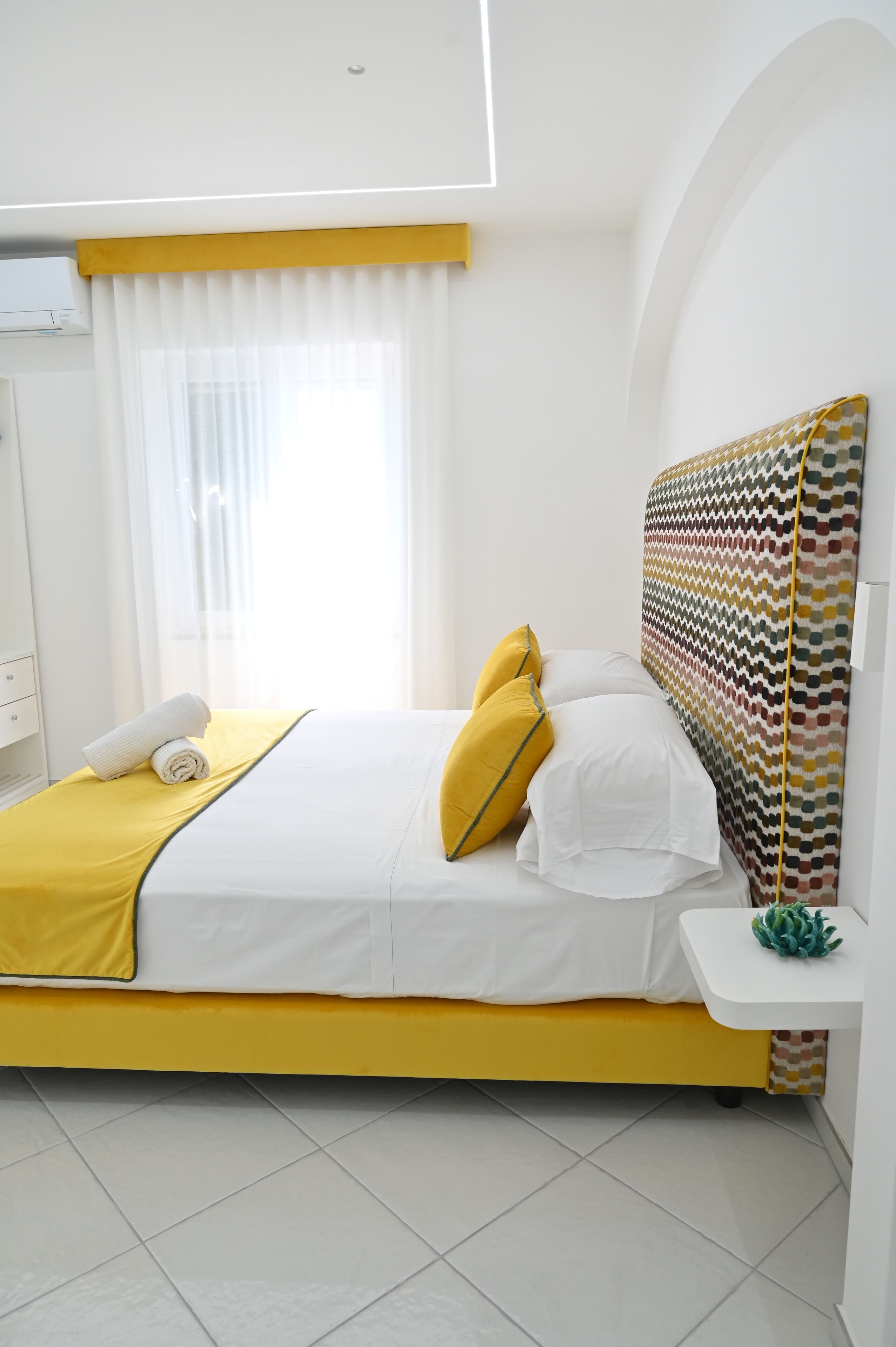 A modern hotel room with a bed, a bedside table, a window with curtains, and a wall-mounted air conditioner.