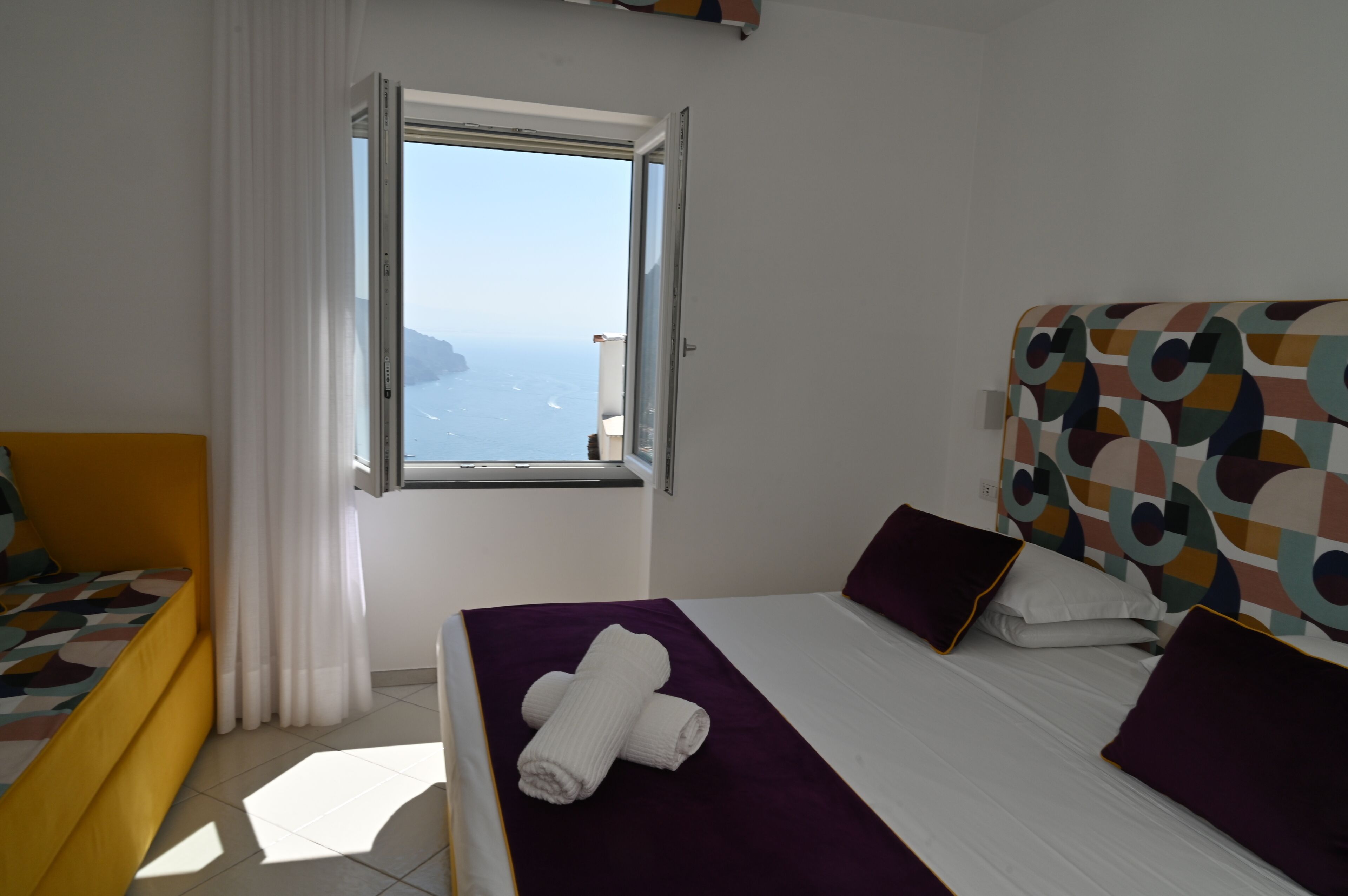 A hotel room with a bed, a window with a view of the sea, and a yellow chair.